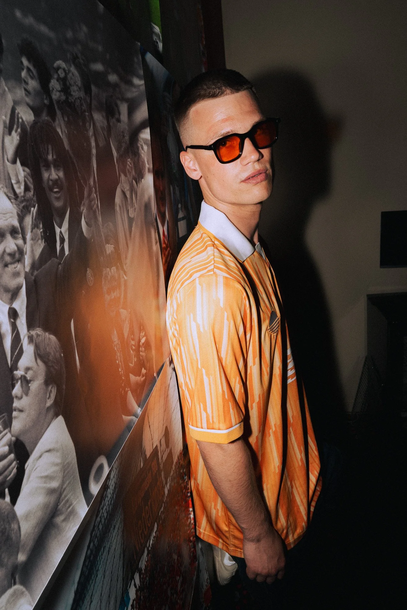 Man met korte broek en oranje en witte shirt, zwart haar en oranje bril, staand naast een muur met zwart-wit foto's van mensen.