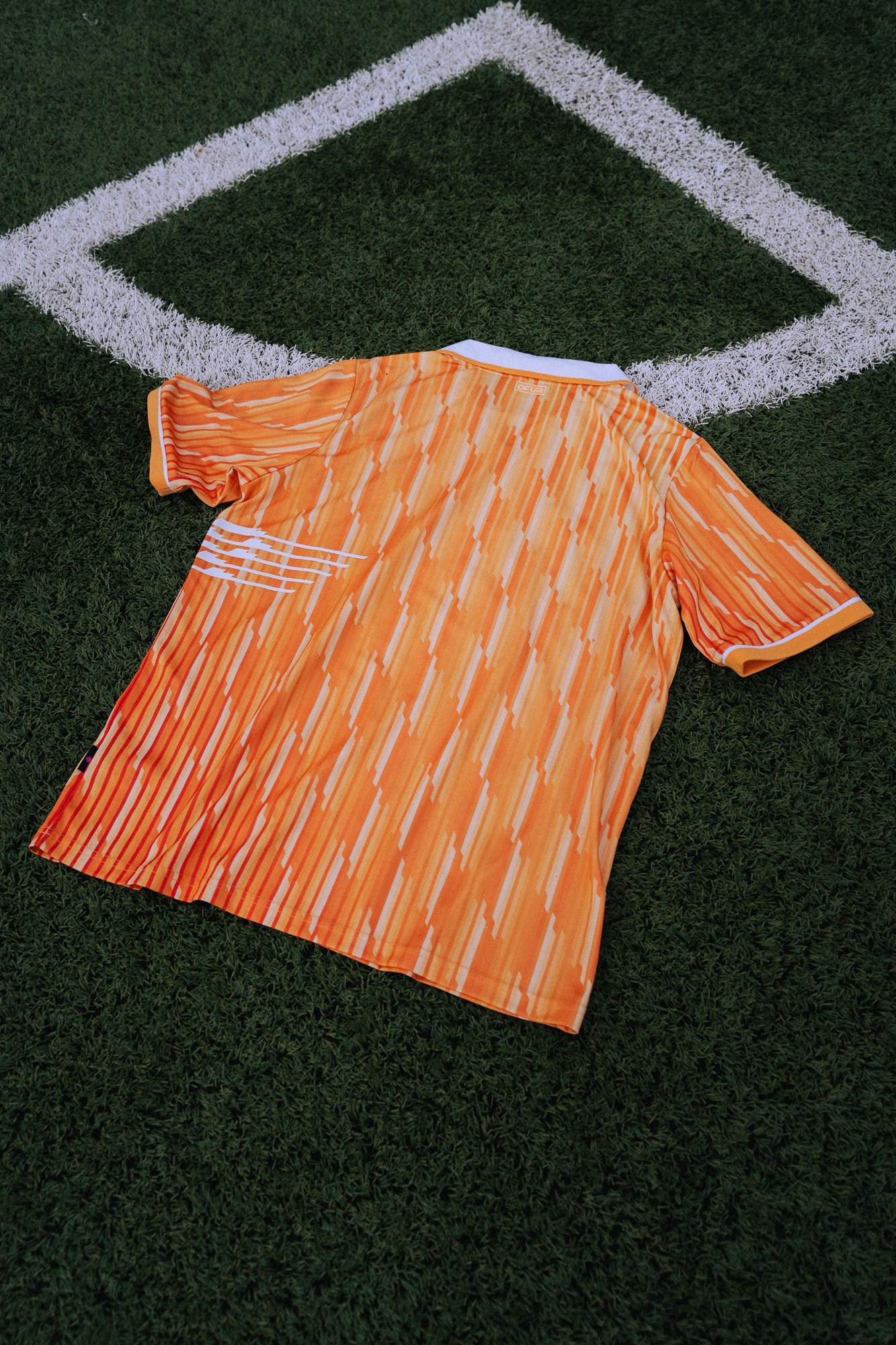 Een oranje voetbalshirt met een patroon van gele strepen ligt op kunstgras, naast een wit uitlijnvlak aan de rand van een voetbalveld.