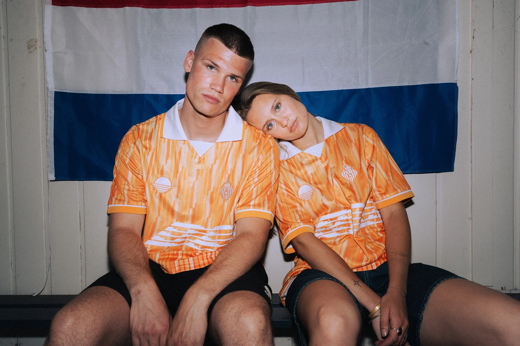 Twee jonge mensen zitten dicht bij elkaar, een man en een vrouw, beiden dragen oranje en wit schaapsjassen met patronen. Ze zitten voor een achtergrond met de vlag van Nederland. Ze lijken ontspannen en casual.