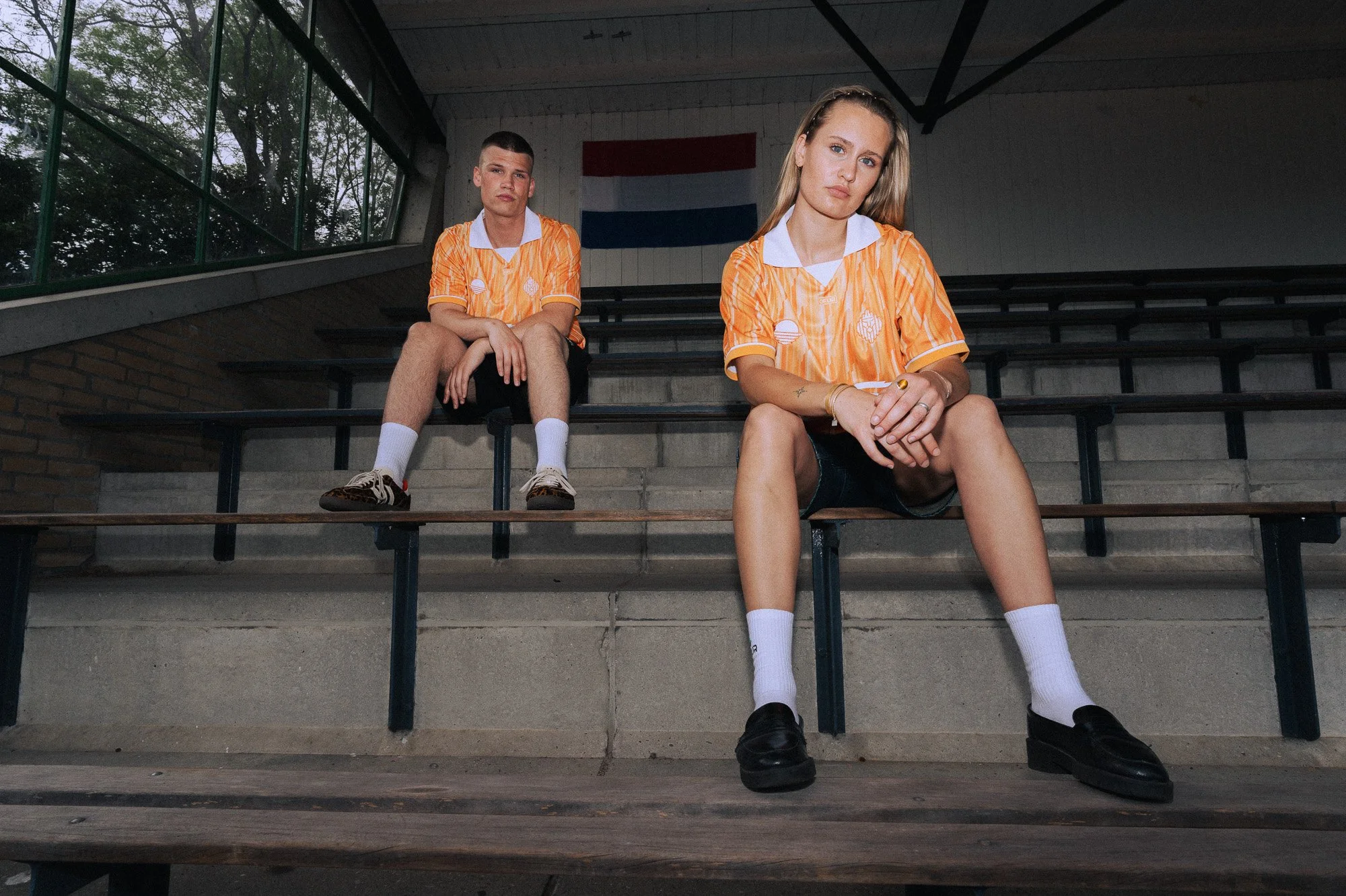 Twee jonge volwassenen in oranje sportshirts zitten op de tribune van een sporthal, met een Nederlands vlag op de muur achter hen.
