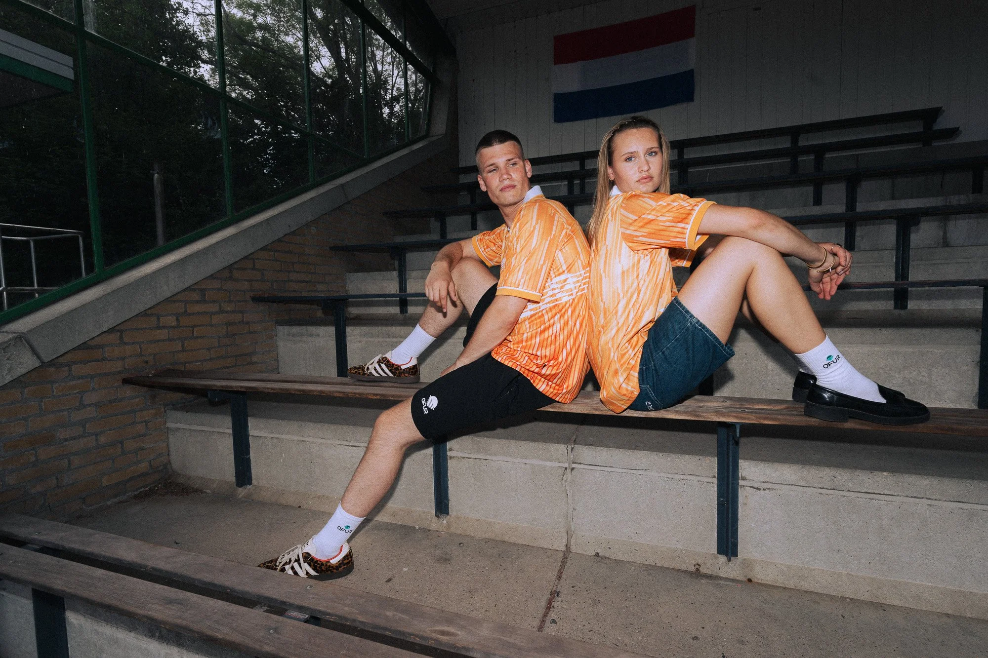 Twee jonge mensen die op een tribune in een sportstadion zitten, gekleed in oranje sportkleding. Ze lijken ontspannen en kijken nieuwsgierig naar de camera.