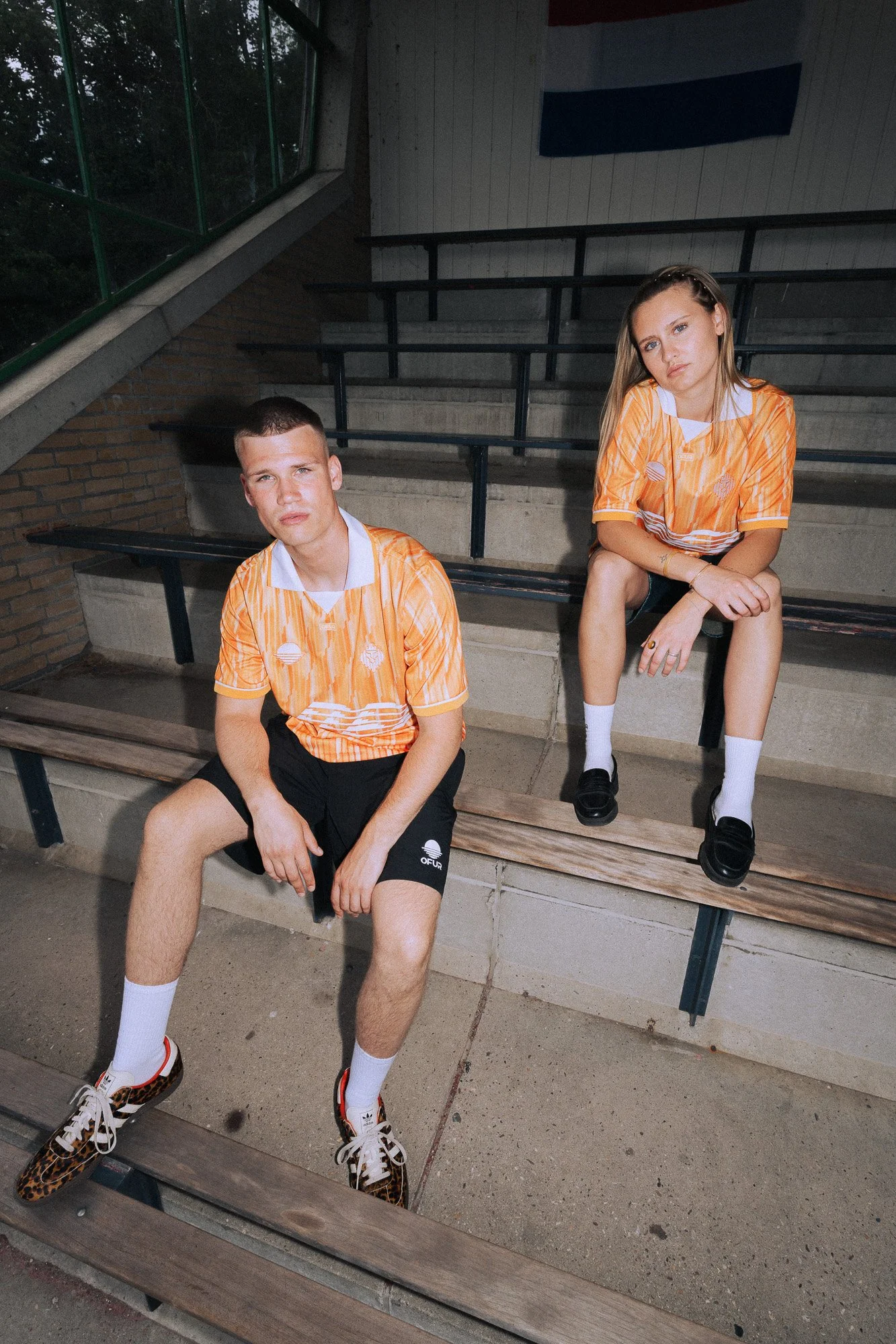 Twee jonge mensen, een man en een vrouw, zitten op een bankje in een buitenomgeving met trappen en ramen op de achtergrond. Ze dragen oranje sportshirts en casual kleding.