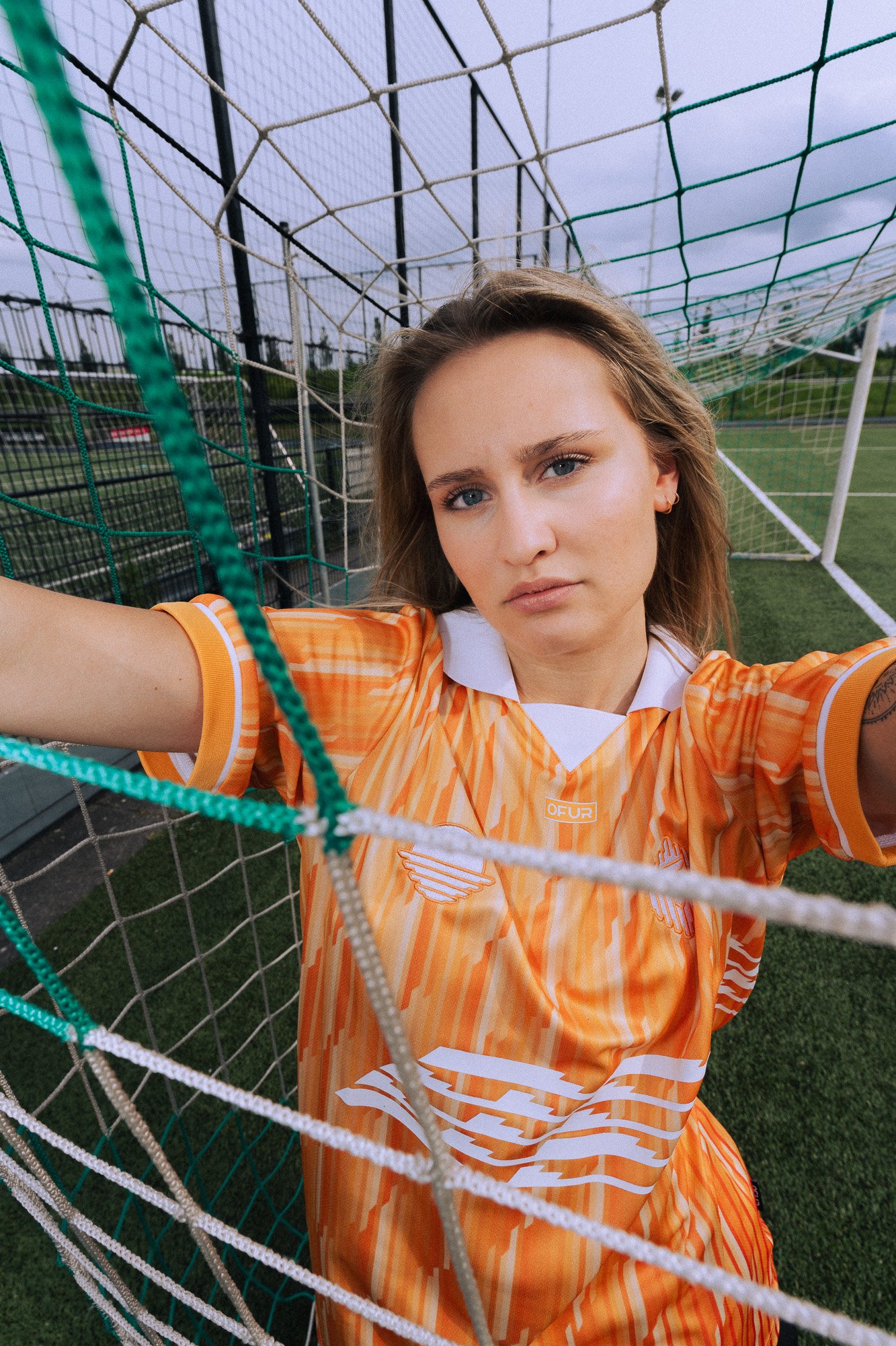 Een jonge vrouw in een oranje en wit sportoutfit neemt een selfie op een sportveld, met het doelnet zichtbaar op de voorgrond.