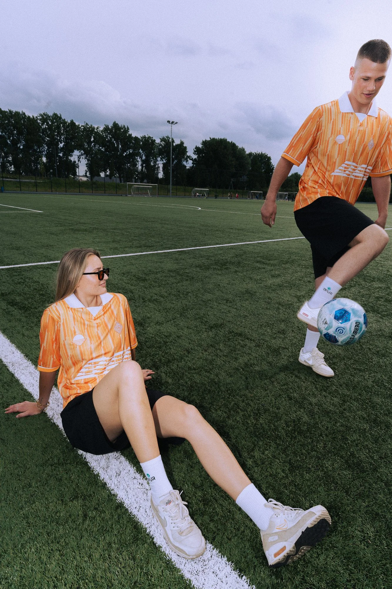 Twee jonge mensen in oranje sportkleding op een grasveld met voetbal, een jongen staat met één voet op de bal en een meisje zit op de grond.