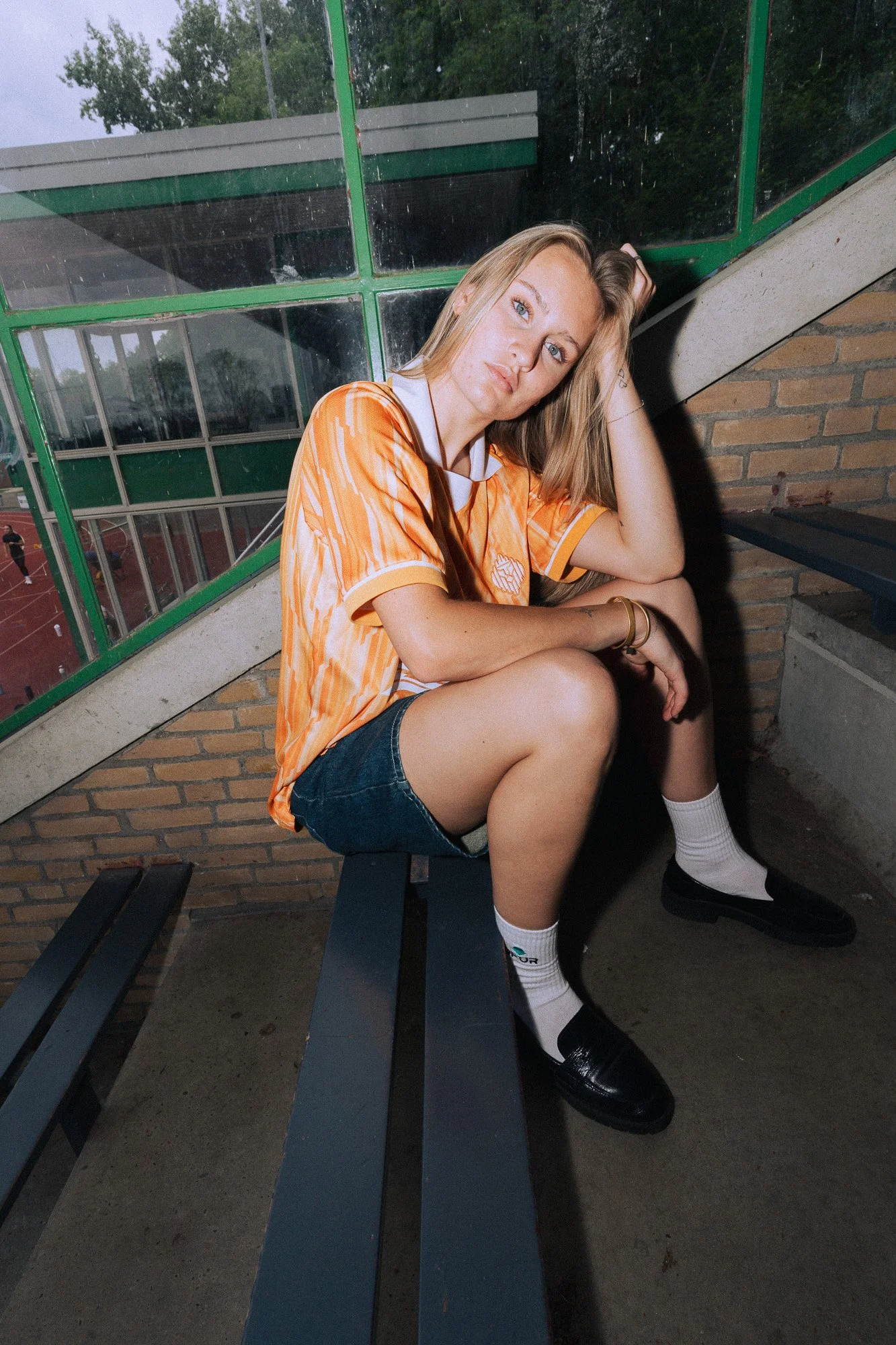 Jonge vrouw met lang blond haar, zittend op een bank buiten, gekleed in een oranje sportshirt en korte broek, met driekleurige sokken en zwarte schoenen.