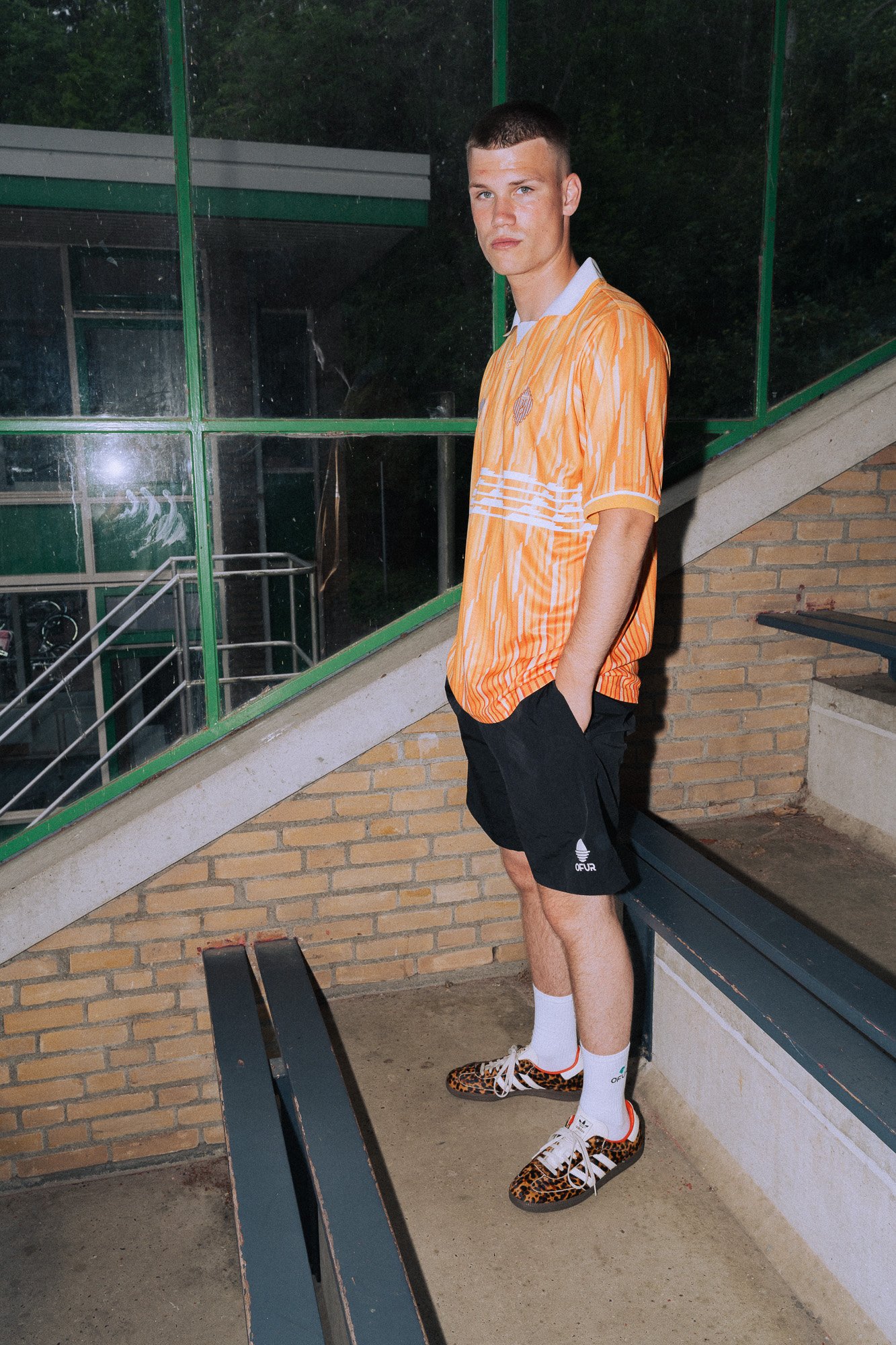 Jongen in een oranje voetbalshirt, zwarte sportshorts, witte sokken en leopardprint schoenen, staand op een bankje naast een stenen muur, met een groene sporthal en glazen ramen op de achtergrond.