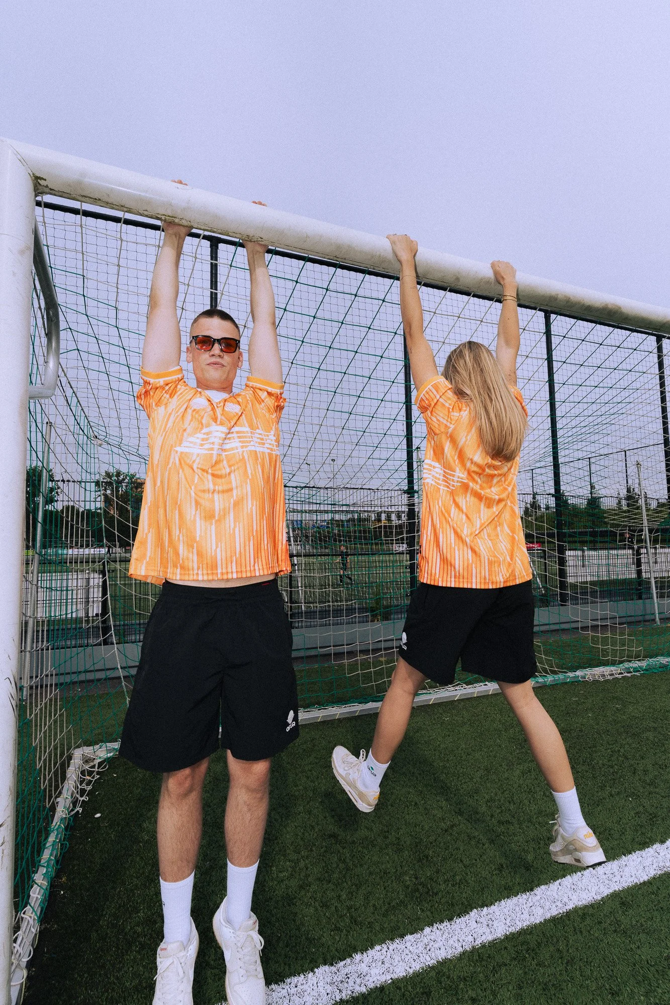 Twee sporters in oranje shirts en zwarte shorts hangen aan een doelpaal op een tennisbaan. Eén draait zich weg, de ander kijkt vooruit met zonnebril.
