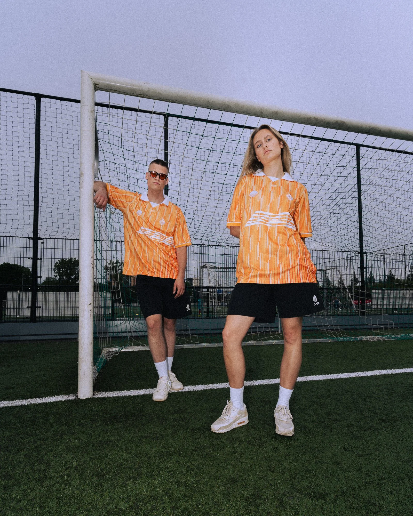 Twee jonge mensen in oranje sportshirts en zwarte shorts poseren onder een voetbaldoel op een kunstgrasveld, met een grijze hemel op de achtergrond.