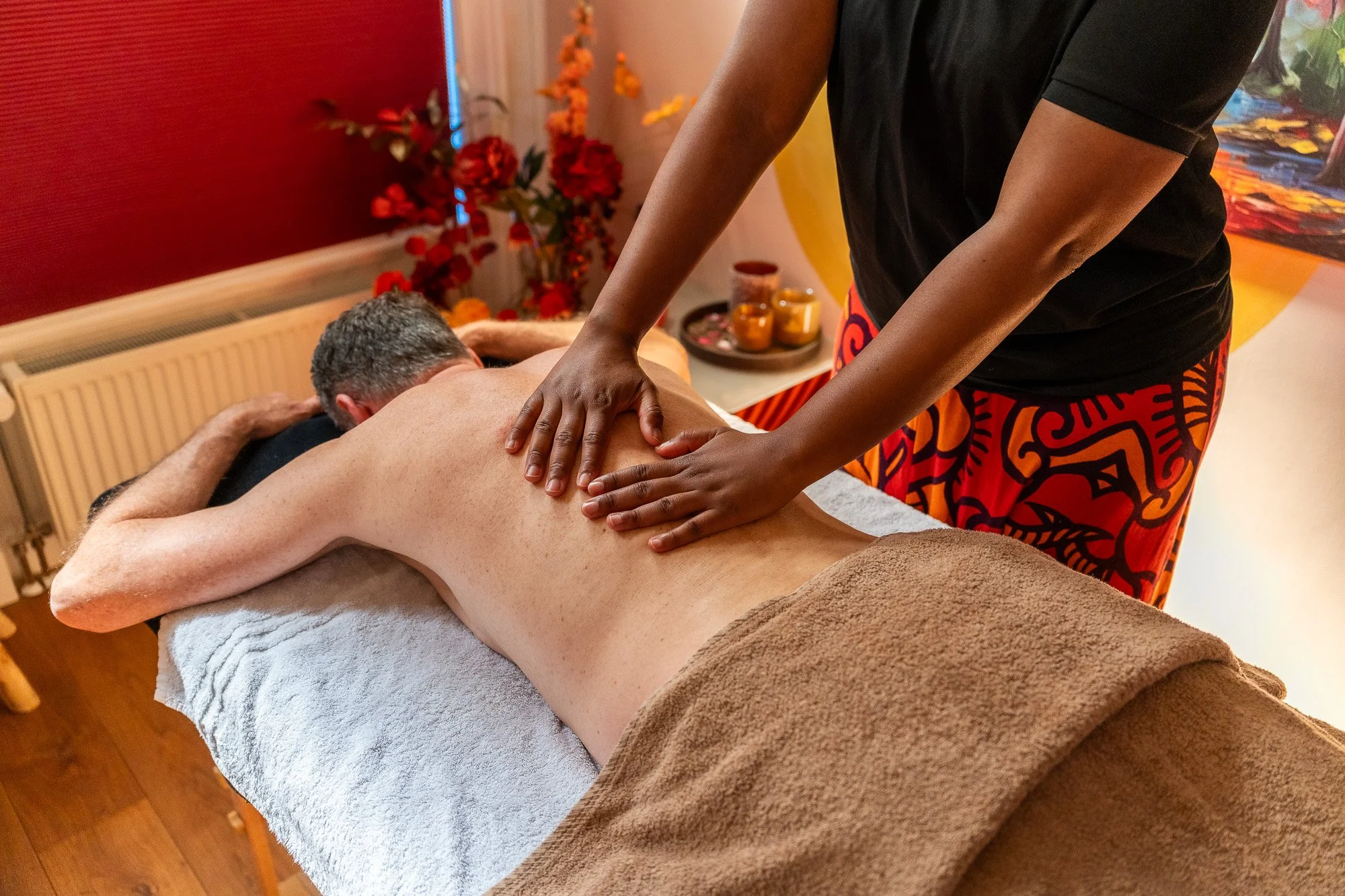 Persoon geeft een massage aan een rug op een massagebank in een ontspanningsruimte met bloemen en decoraties op de achtergrond.