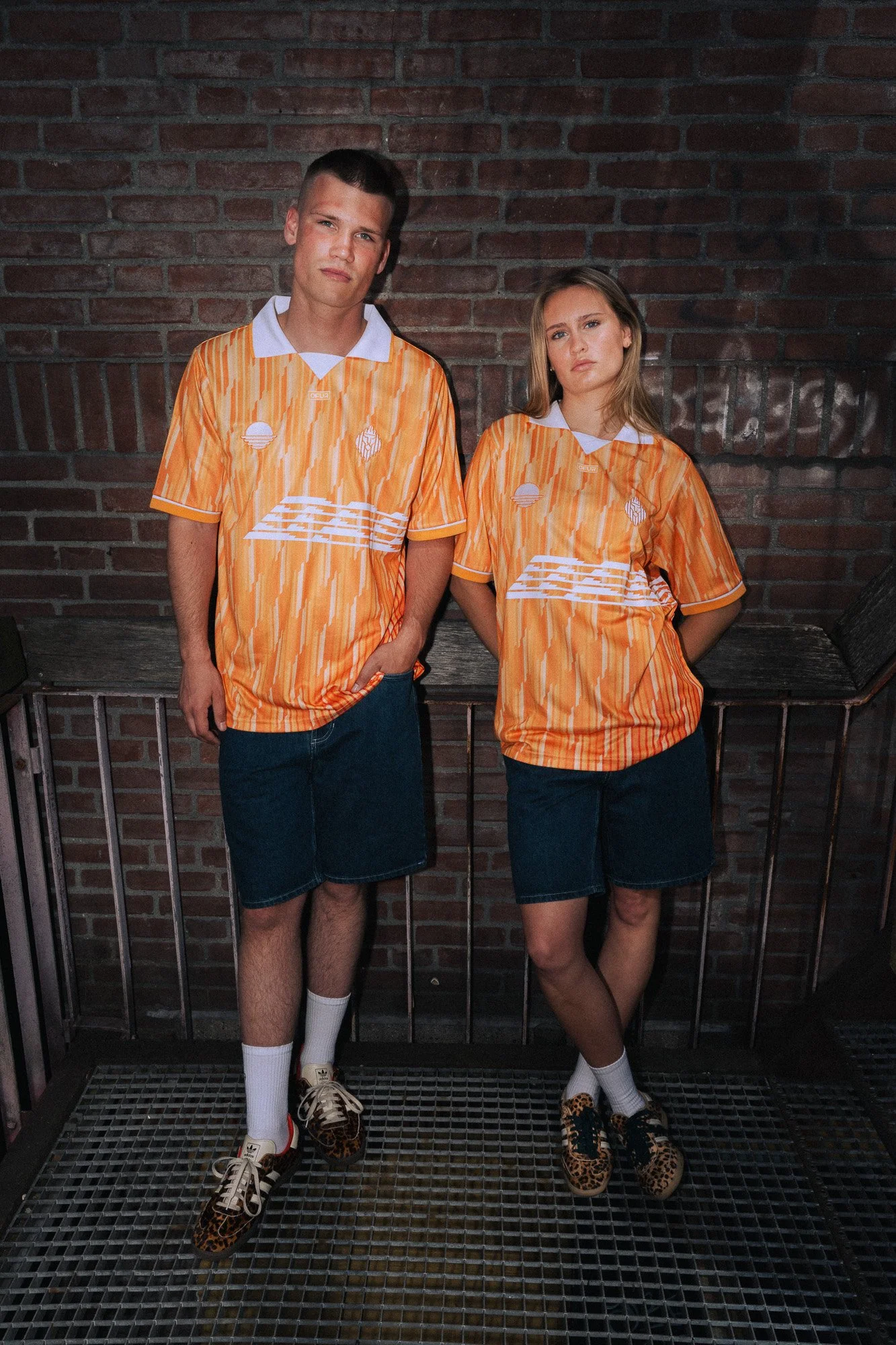 Twee jonge mensen dragen gelijkaardige oranje sportshirts met wit en grafische patronen, staan voor een bakstenen muur, met een metalen hek erachter.
