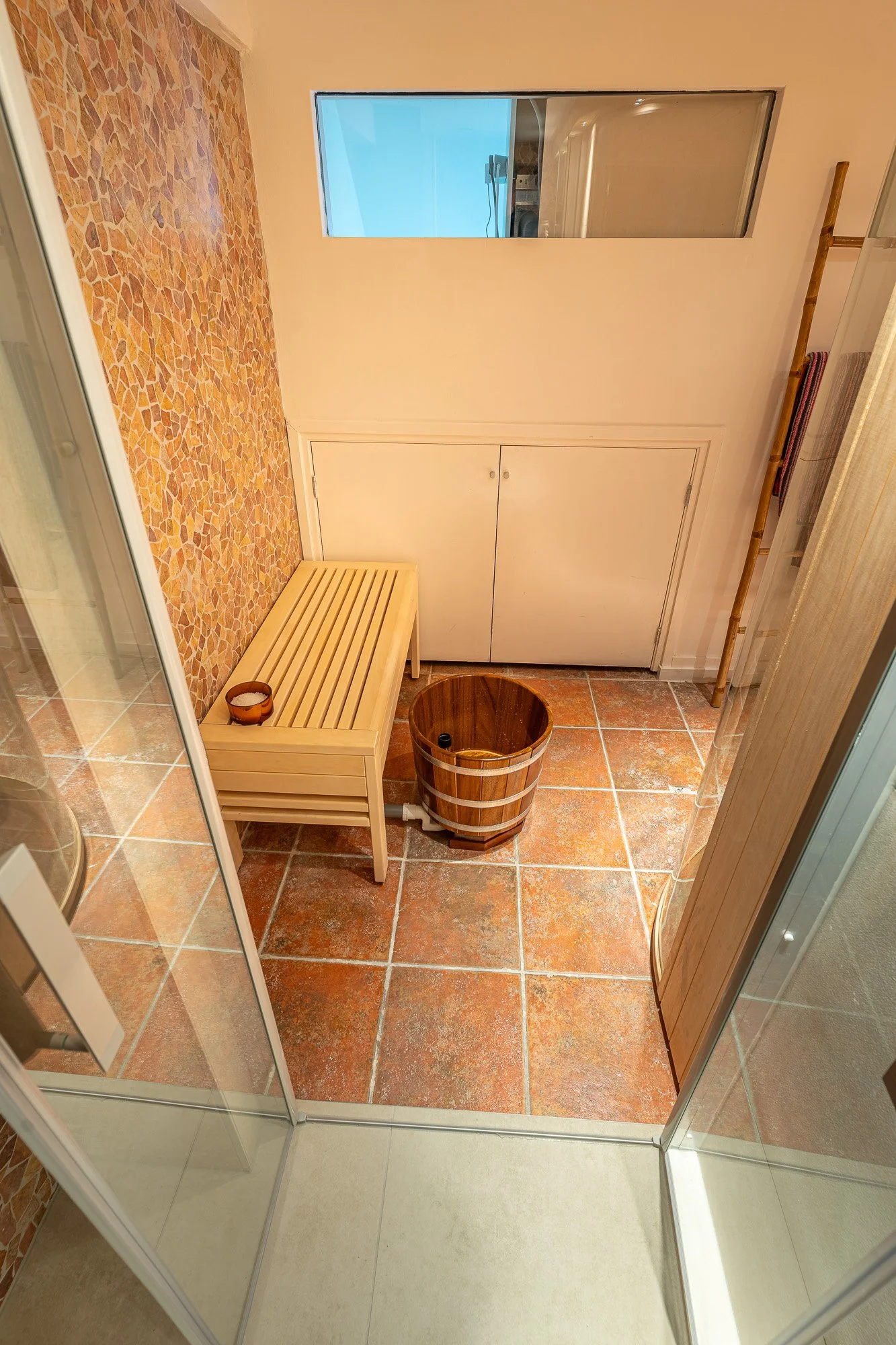 De foto toont een separaat sauna- of wellnessruimte met een houten banken, een houten emmer, een spiegel, en een bamboe stok met een doek. De vloer is betegeld met terracotta-kleurige tegels. Er is een witte kast en een raam dat uitkijkt op een ruimt