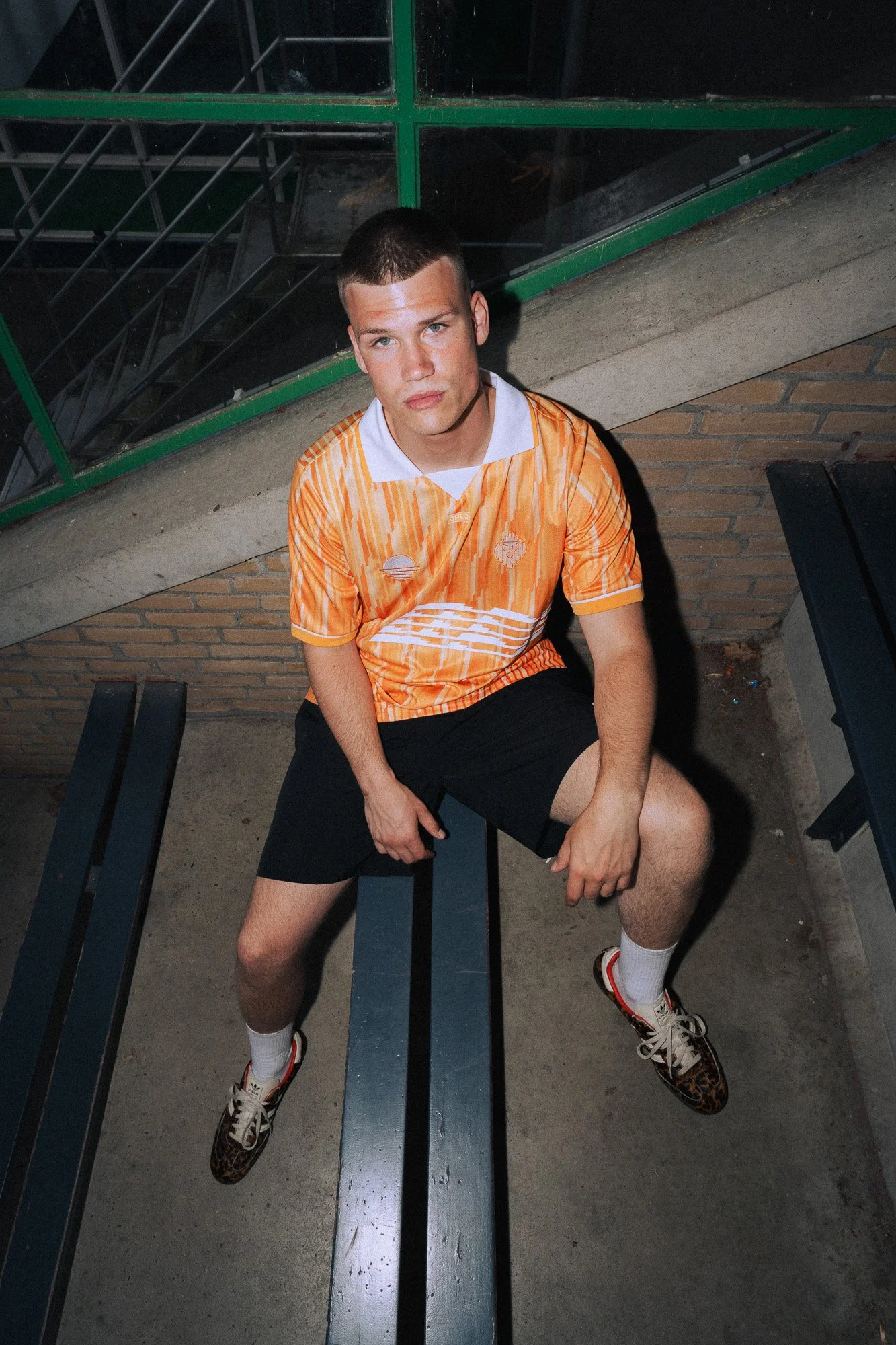 Een jonge man zit op een bankje buiten, gekleed in een oranje gestreept voetbalshirt, zwarte korte broek, witte sokken en sneakers met luipaardprint.