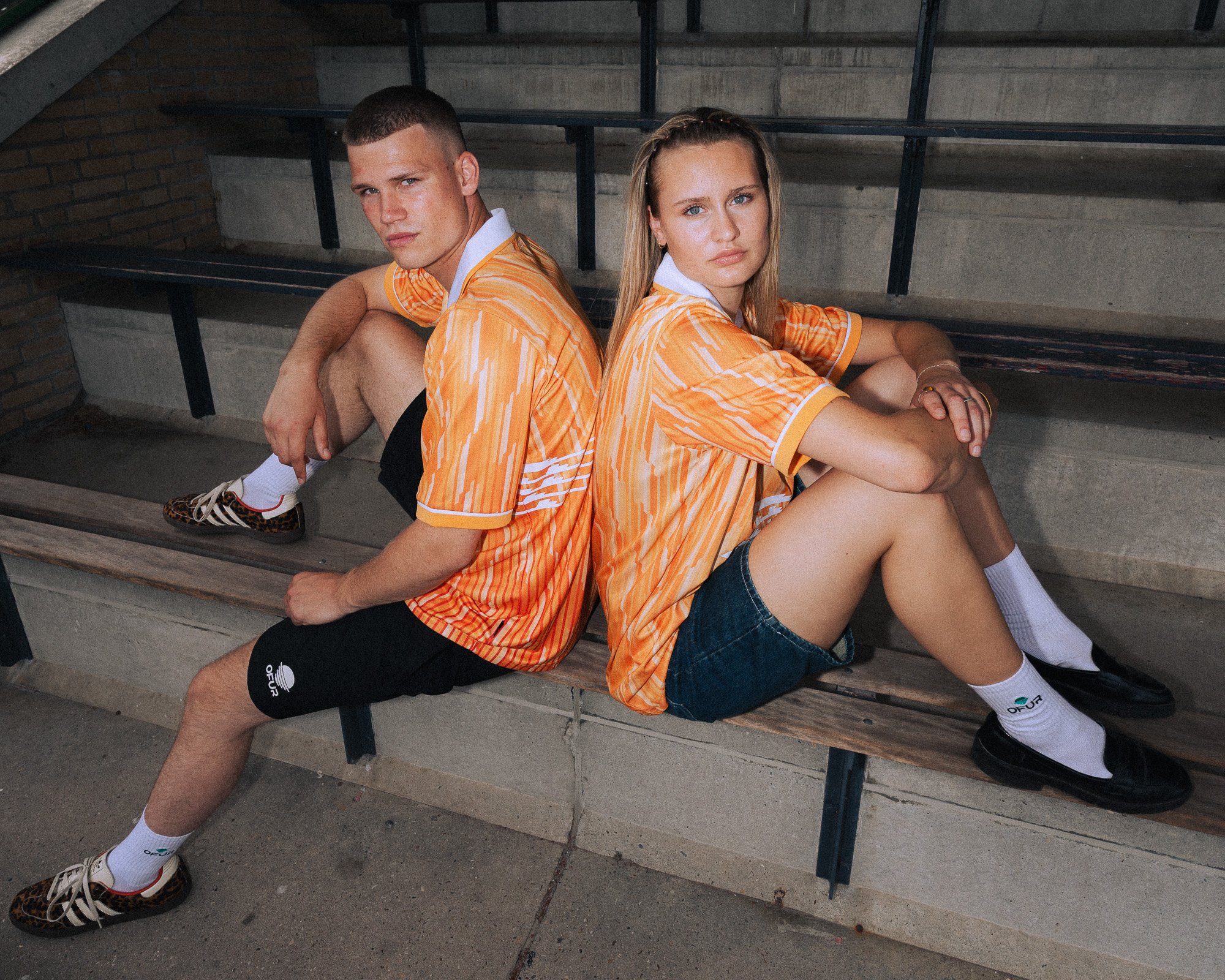 Twee jonge mensen zitten op een bankje onder een betonnen trap, gekleed in oranje sportkleding, met een strakke haardracht en diverse sieraden.