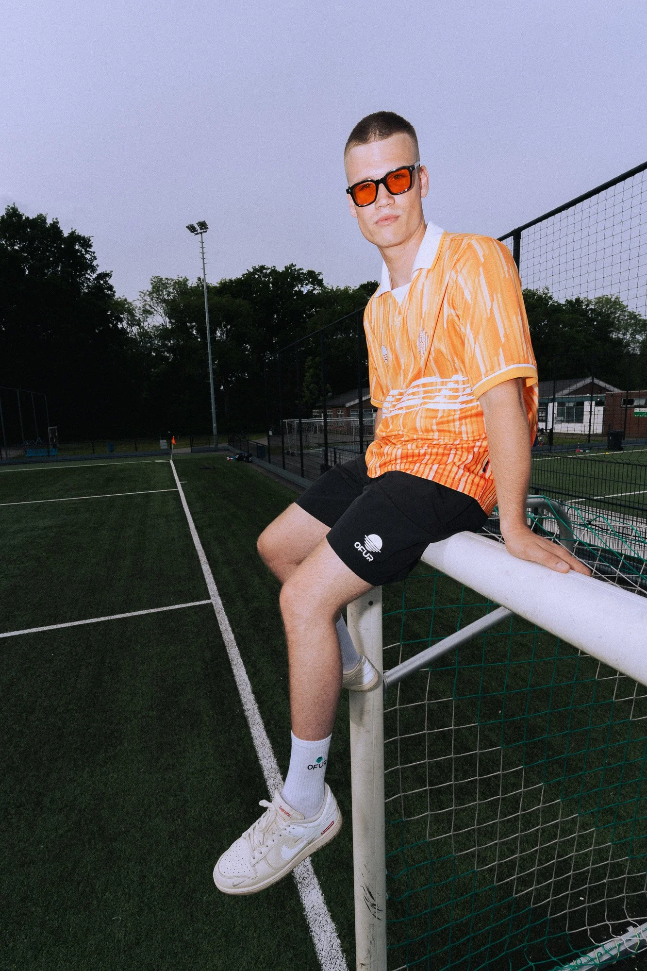 Jongen zittend op een voetbalveld naast een sportdoel, draagt een oranje en witte shirt, zwarte shorts, witte sneakers en donkere zonnebril, met een achtergrond van bomen en een lichtmast.