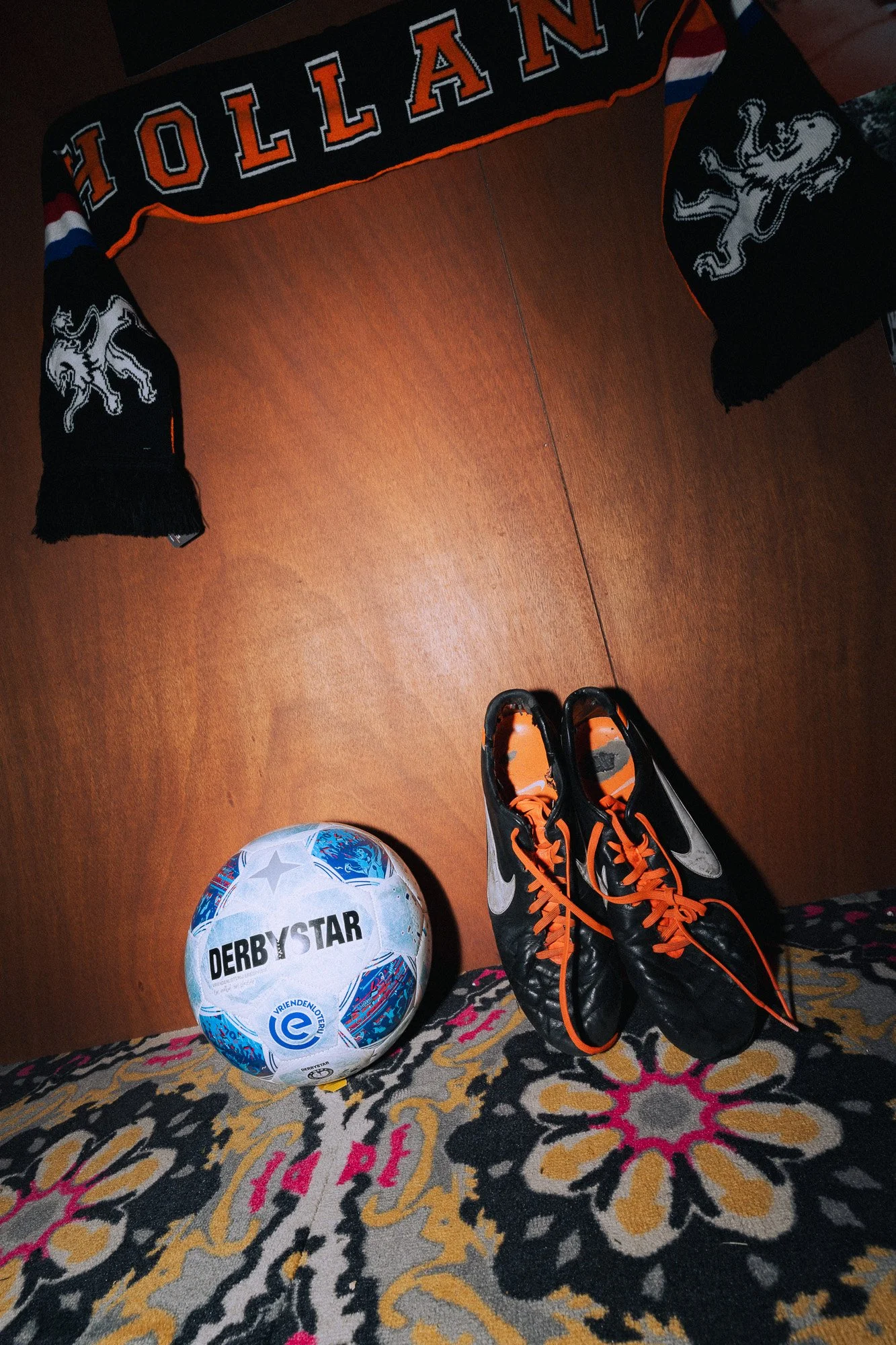 Een bal met de tekst 'DERBYSTAR' op een tapijt, een paar zwart en oranje sport sneakers, een voetbal en een gescheiden wollen sjaal met de naam 'HOLLAND'}