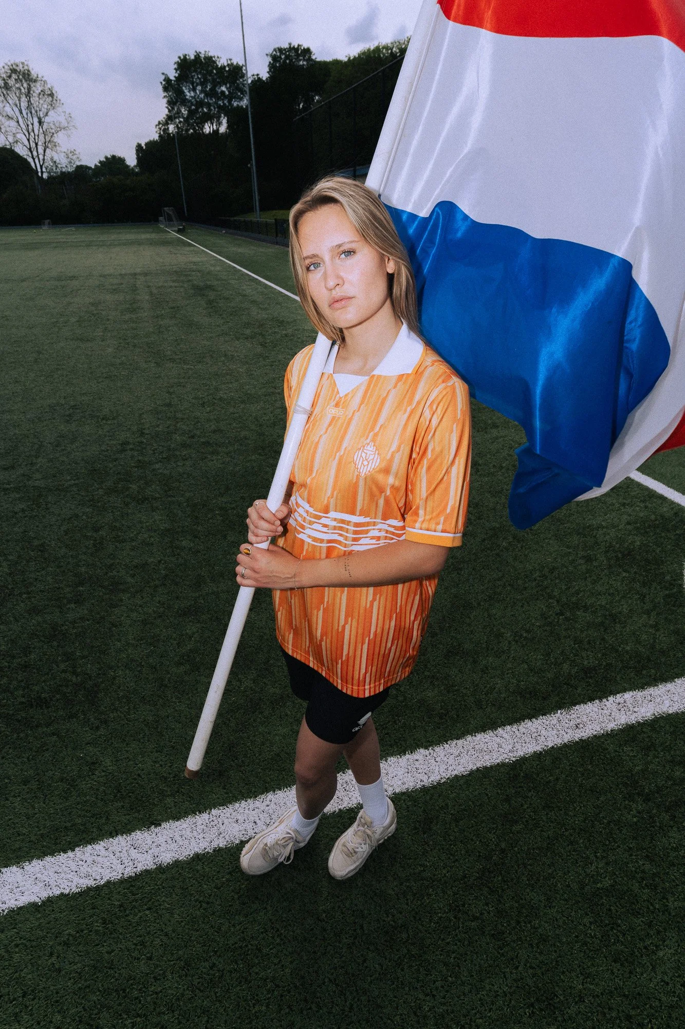 Een jonge vrouw houdt een vlag met de kleuren rood, wit, en blauw op een voetbalveld. Ze draagt een oranje sportshirt, zwarte sportbroek, witte sokken en schoenen.