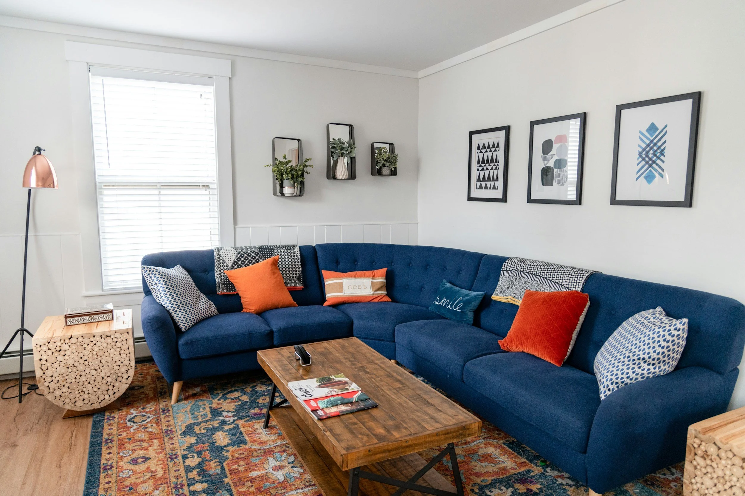 Living room with a blue sectional sofa decorated with orange, gray, and patterned pillows, a wooden coffee table, a floor lamp, wall art, and a window with blinds.