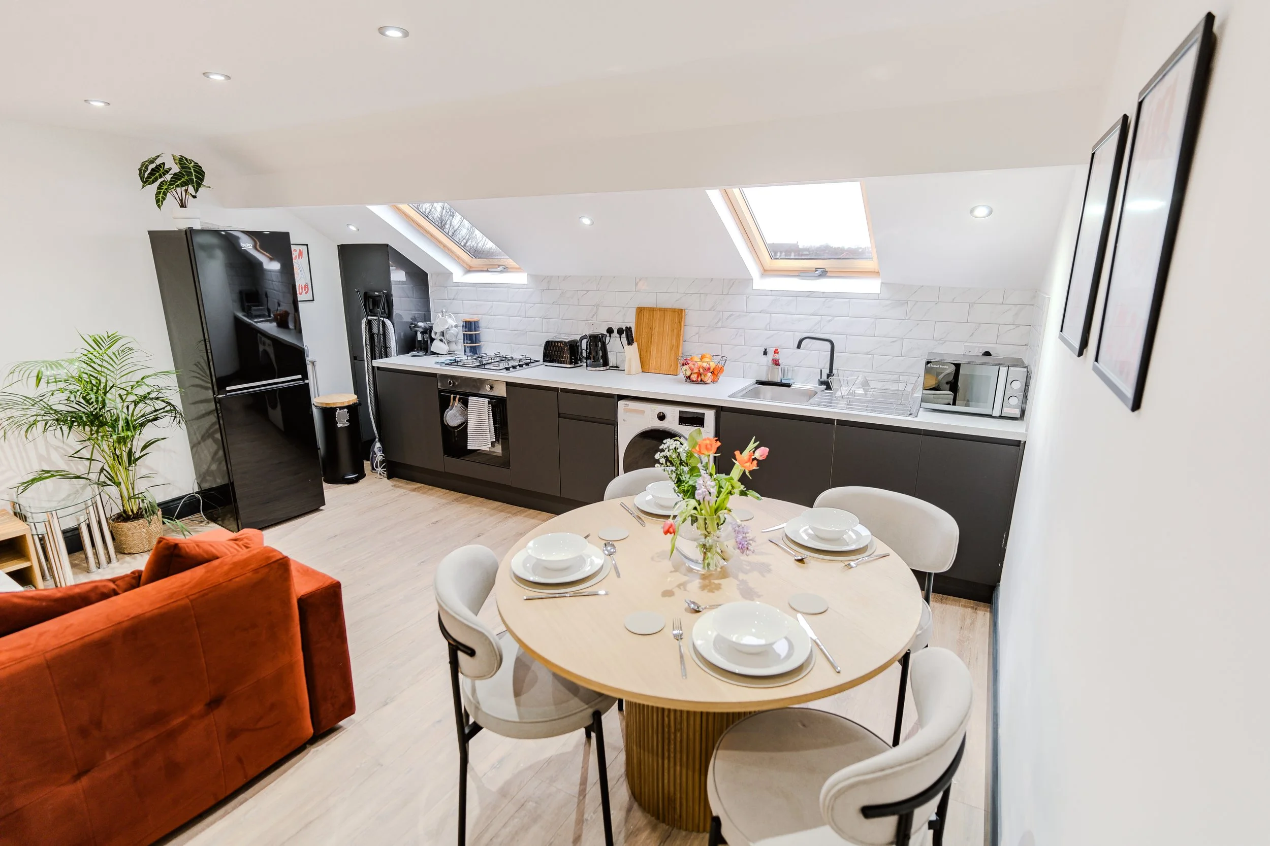 Modern kitchen with black cabinetry, white countertop, two skylights, and white brick backsplash, adjacent to a dining table set with plates and a floral centerpiece.