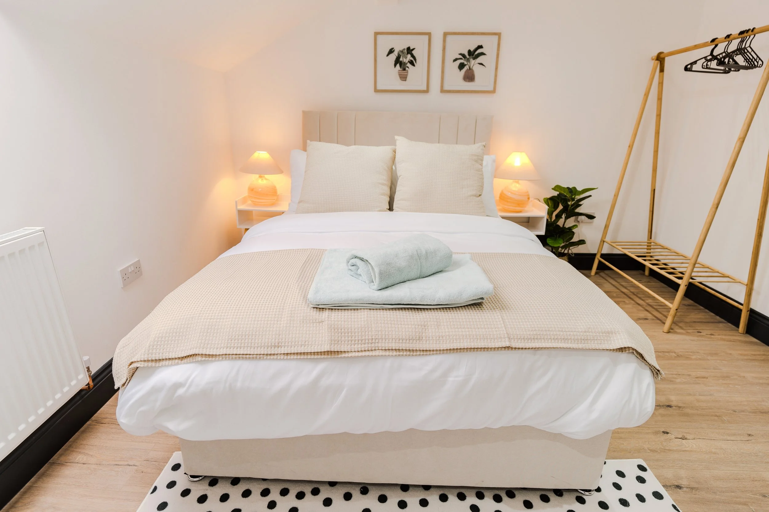 A neatly made bed with three pillows, two of which are beige and one white, in a bright bedroom with two lamps on nightstands, framed plant artwork above, a beige blanket, a rolled towel with a matching blanket on top, a wooden clothing rack with han
