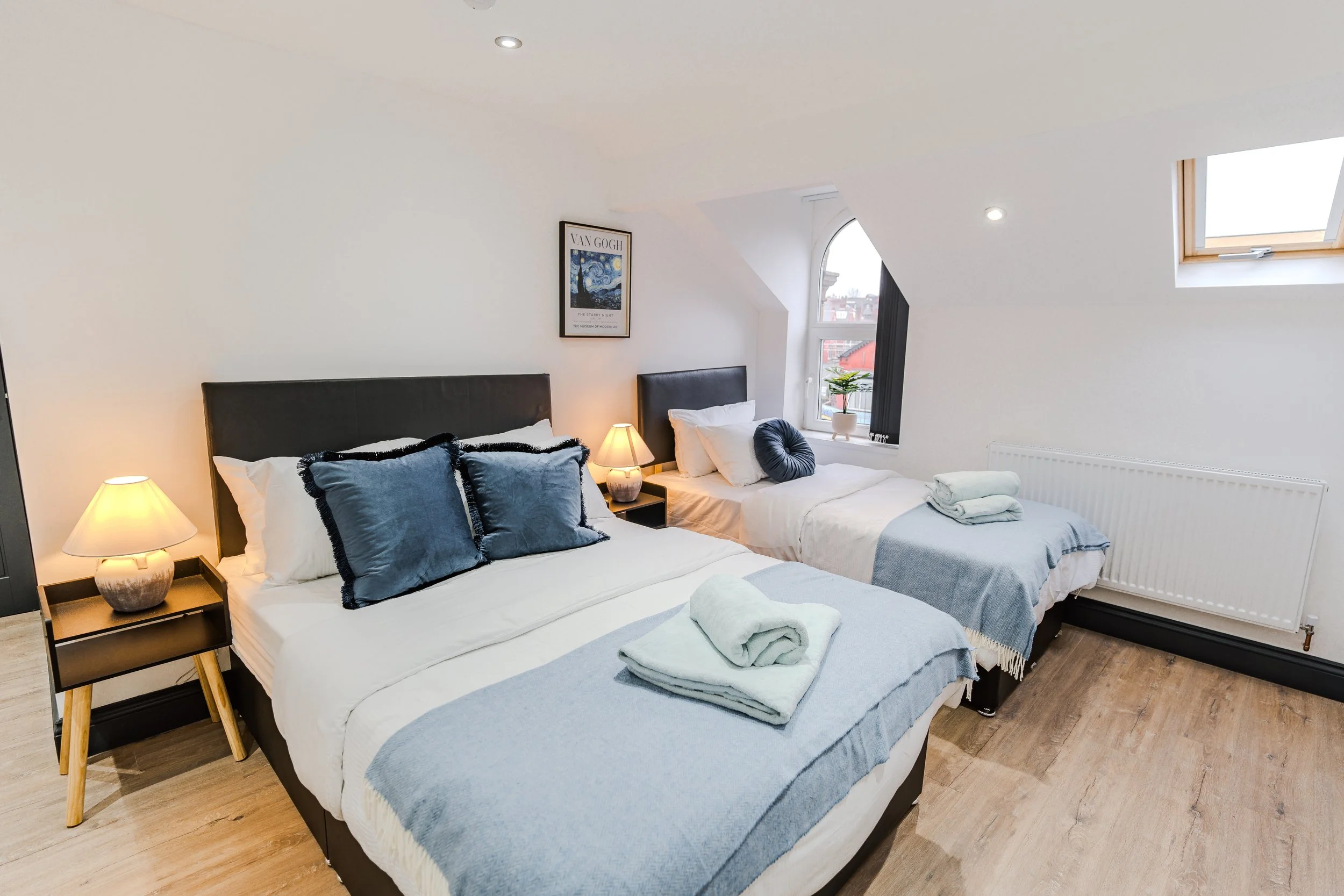 A bright bedroom with two beds, each with white linens and blue pillows. Small nightstands with lamps sit between the beds. Towels are folded on the beds, and a window with a potted plant is visible. The room has wooden floors and a sloped ceiling with skylights.