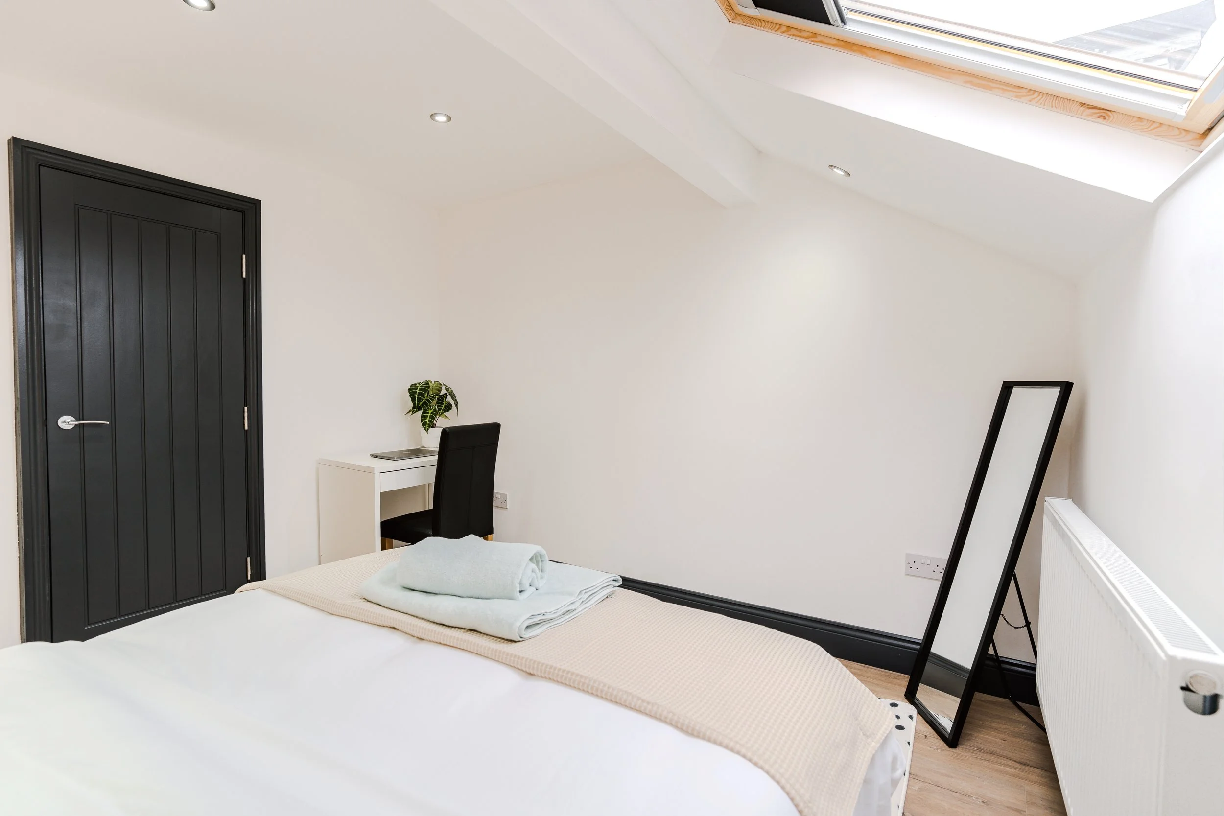 A minimalist bedroom with a white bed, folded towels on top, a black door, a small desk with a chair, a plant, a leaning mirror, and a skylight window letting natural light in.