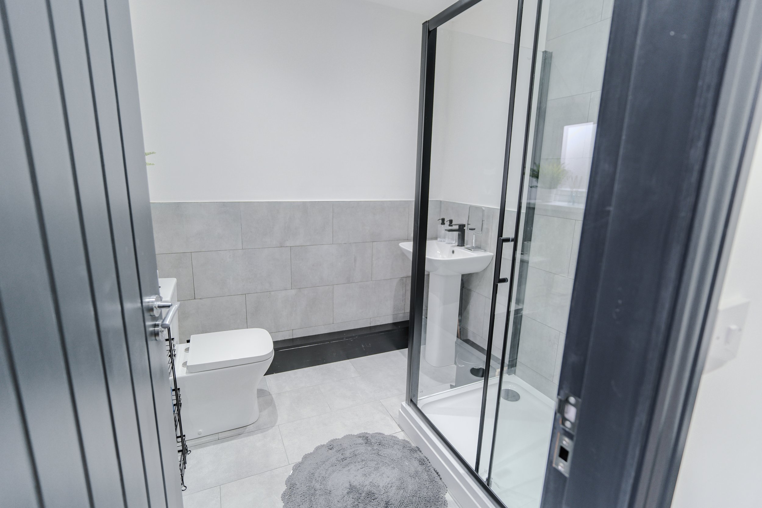 View of a modern bathroom with a toilet, a sink with a mirror, a glass shower enclosure, a round gray rug, and gray tiled walls and floor.