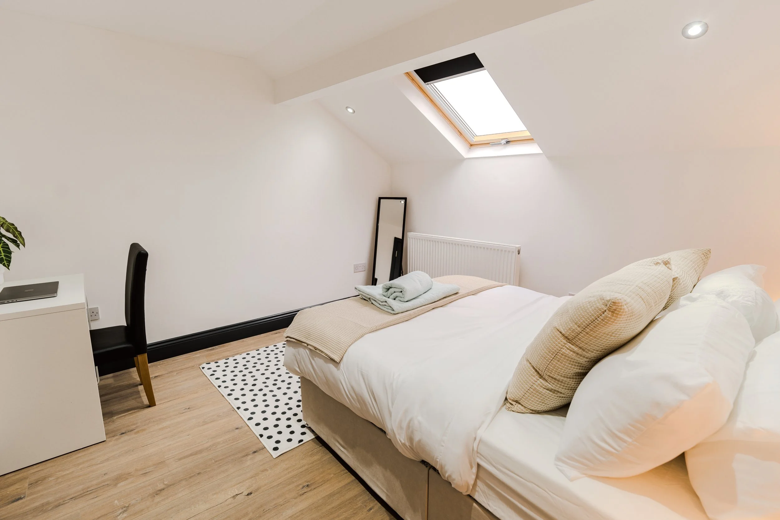 A tidy bedroom with a bed, pillows, towels, a mirror, a desk, and a skylight window.