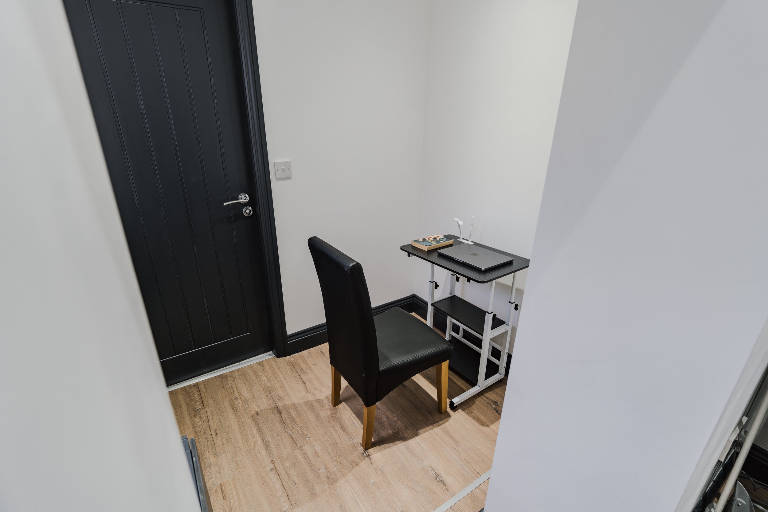 A small corner of a room with a black door, a black chair, a small black and white desk with a laptop, a book, and a lamp on top.