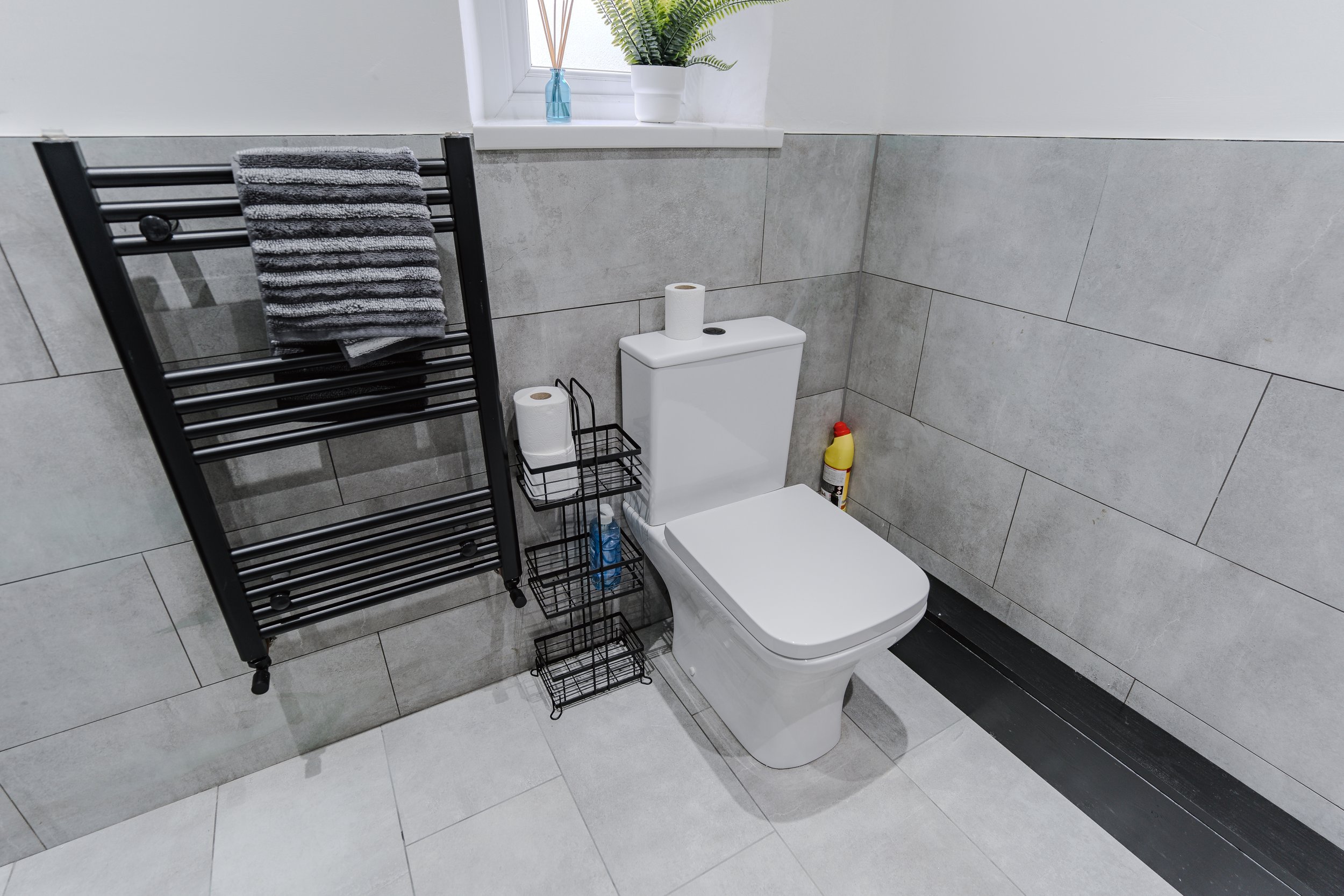 Modern bathroom with a white toilet, black towel rack with gray towels, potted plant on window sill, toilet paper on holder, cleaning spray behind the toilet, and a window with natural light.