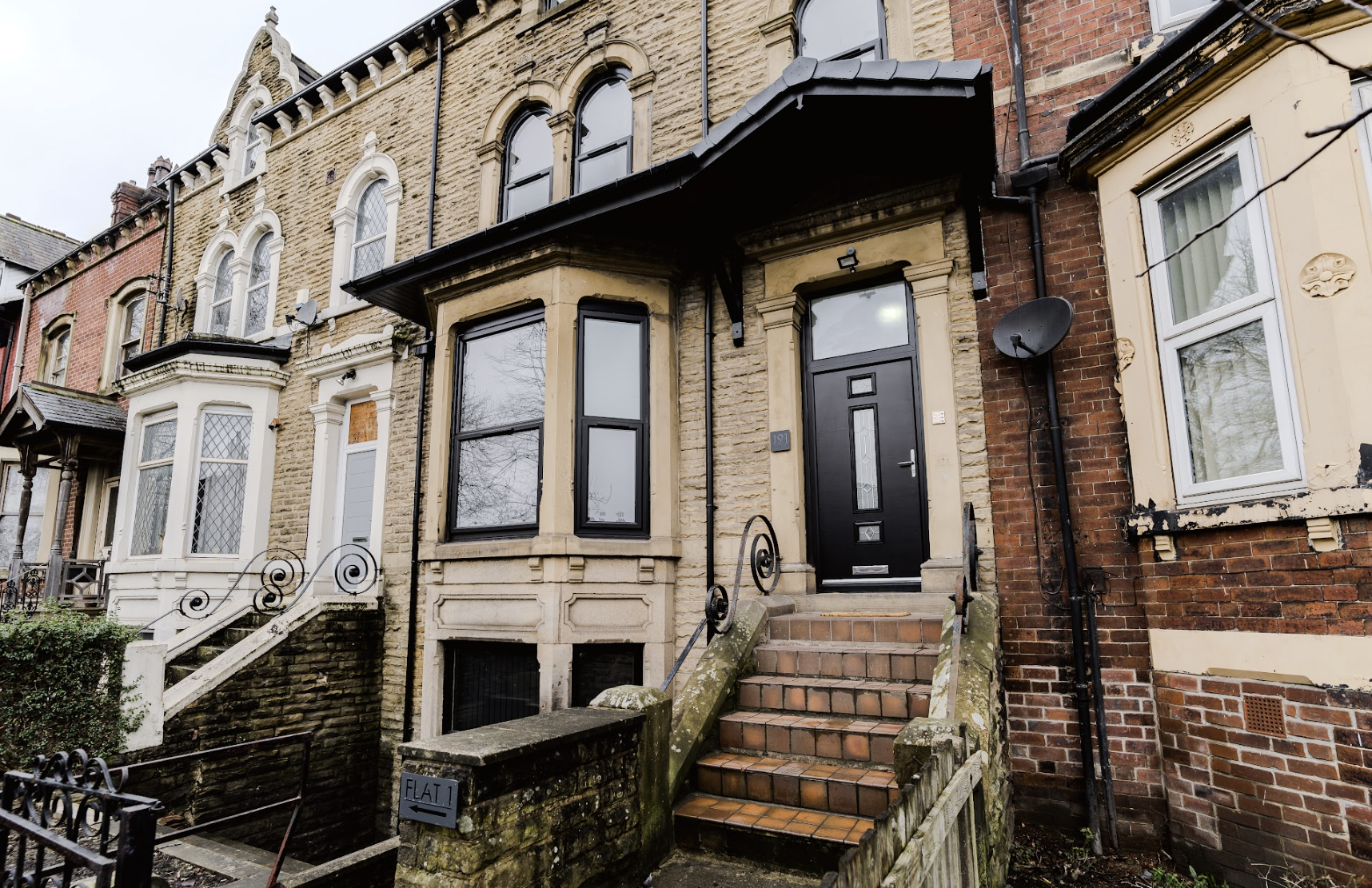 Front view of a row house with a black front door, steps, and a bay window, located on a street with other similar houses.
