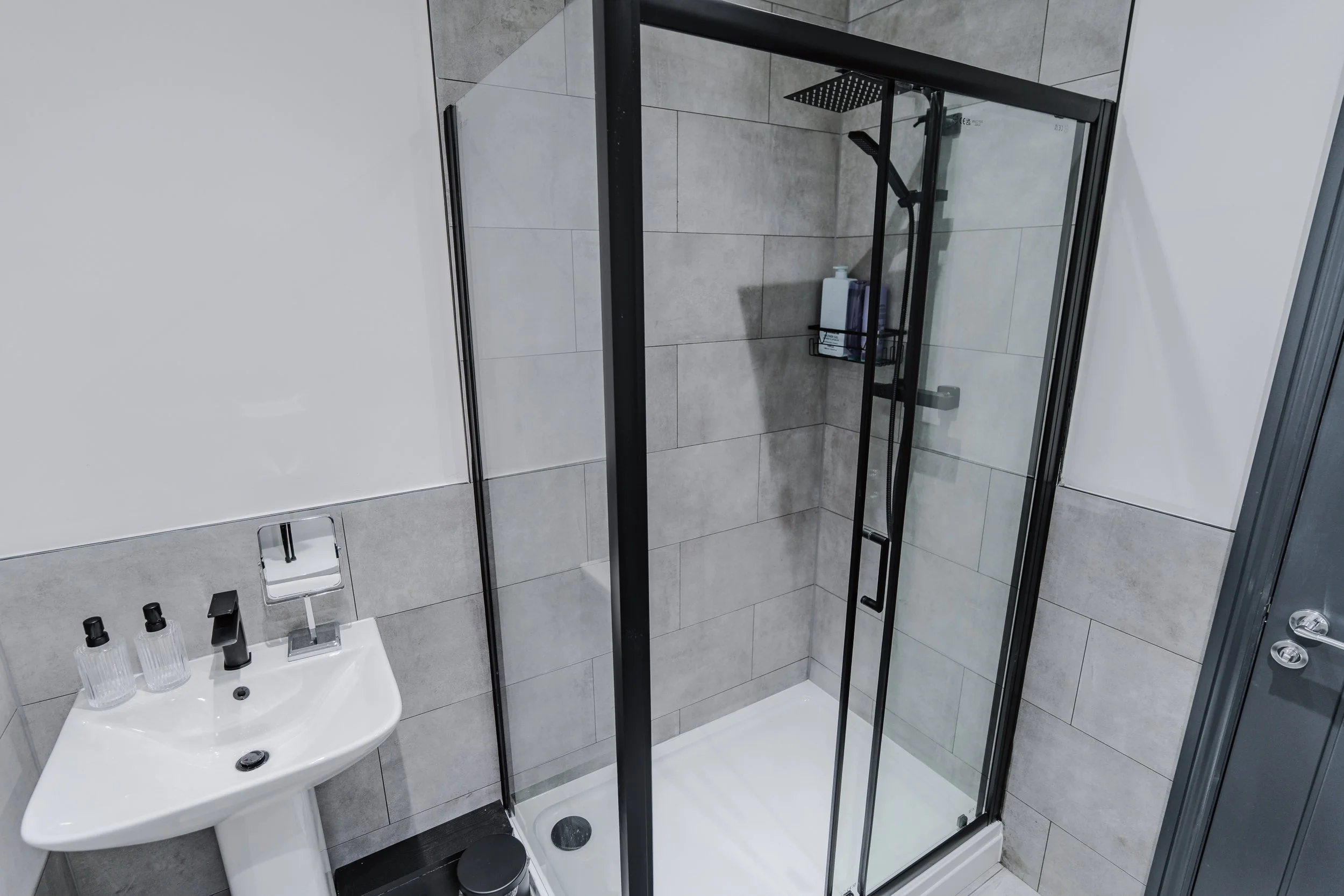Modern bathroom shower with glass doors, gray tiled walls, and a black shower fixture. There are bottles of toiletries on a black shelf inside the shower. A small white sink with a black soap dispenser and bottles on top is adjacent to the shower.