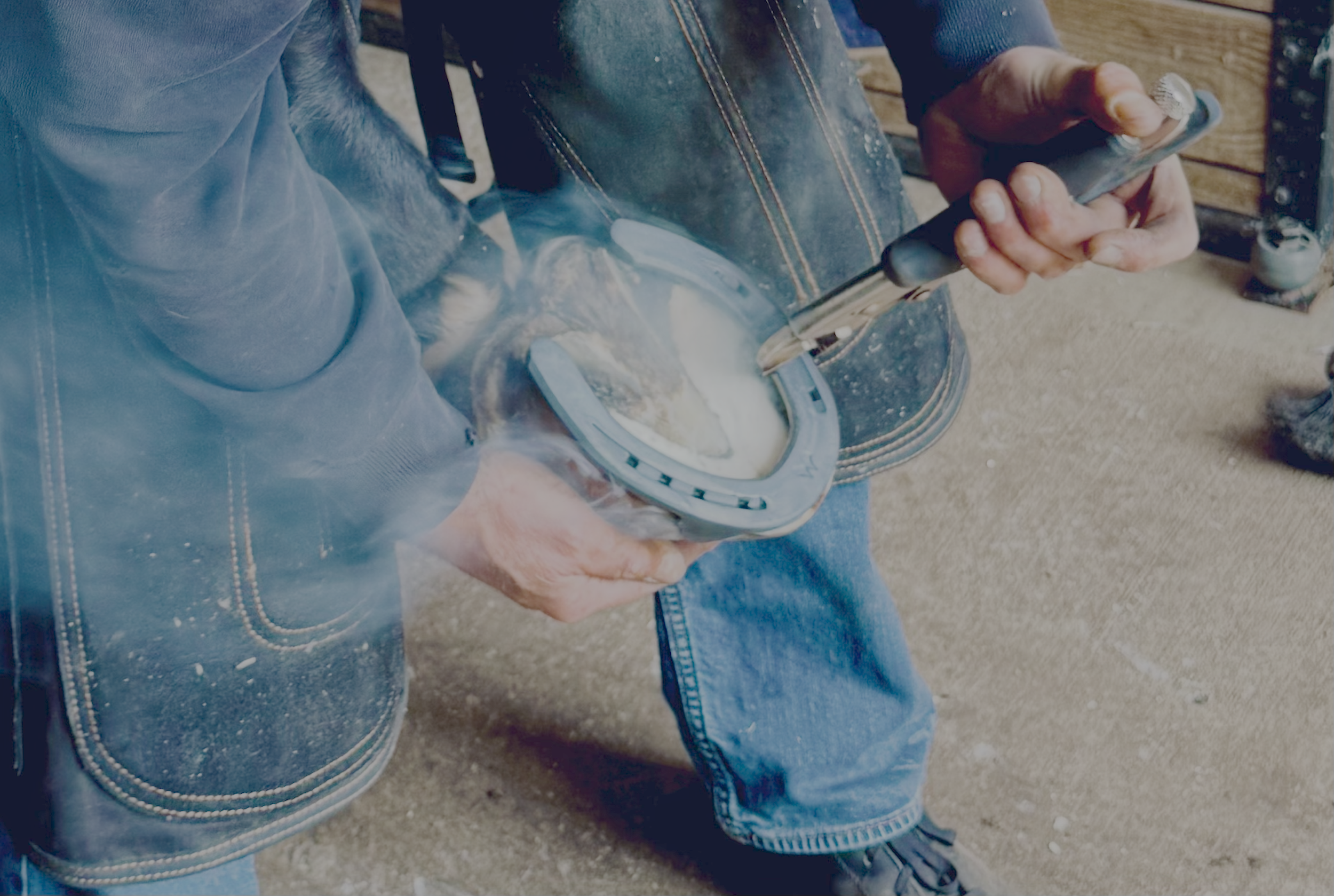 Person in blue jeans and a dark jacket holding a smartphone while working on a horseshoe on a workbench, with a hammer nearby.