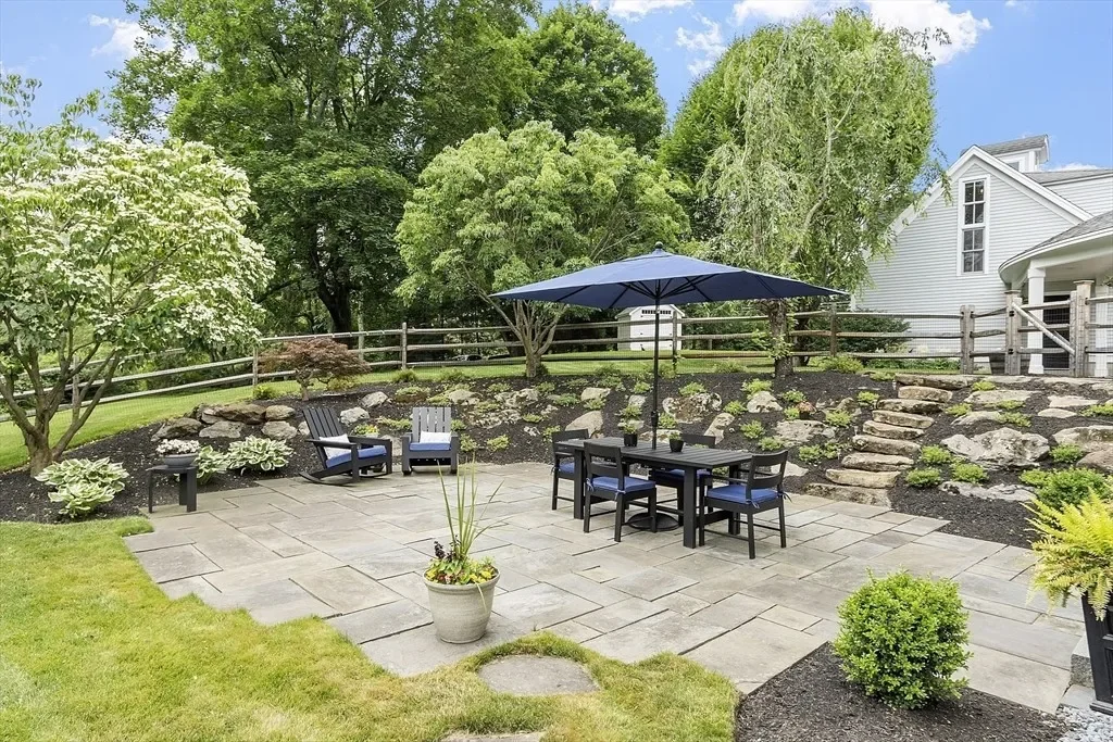 A backyard patio with outdoor furniture, including a table with an umbrella, surrounded by greenery and a stone retaining wall with steps.