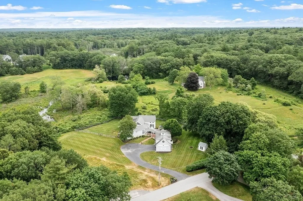Aerial view of a large countryside property with a white house, garage, driveway, and extensive green lawn surrounded by trees, fields, and forest.