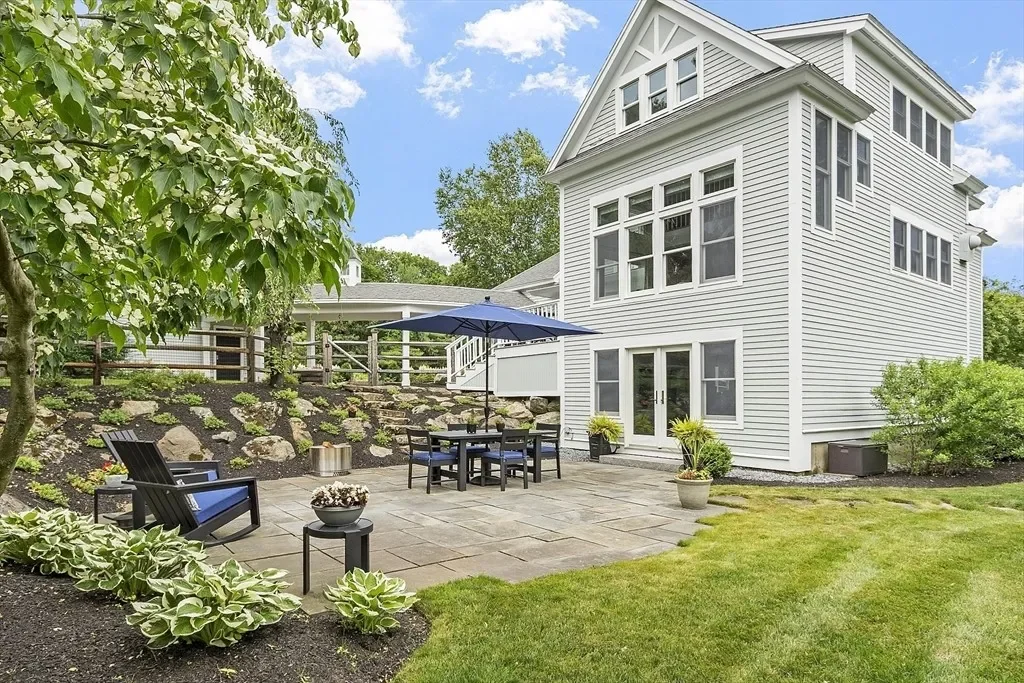 Backyard patio area with outdoor furniture, including a table with a patio umbrella, two chairs, and potted plants, with a white house in the background and lush greenery.