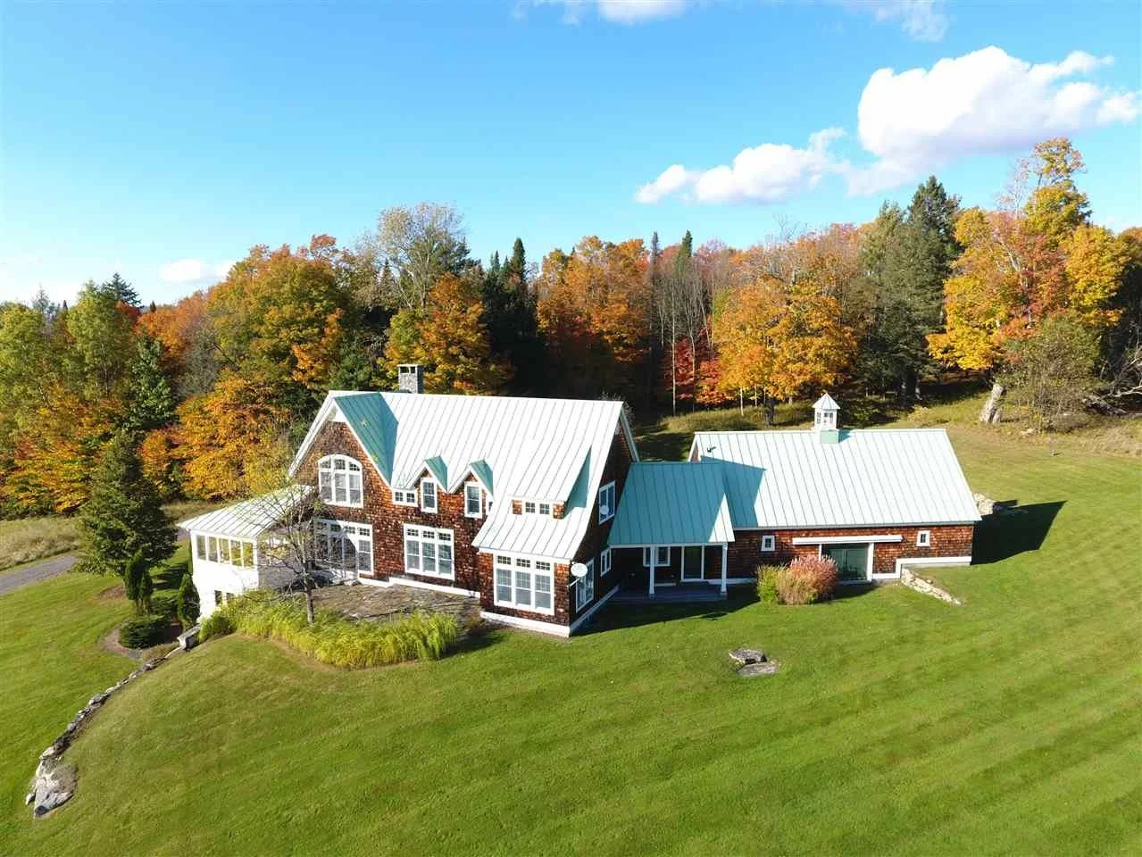 A house with a metal roof surrounded by green lawn and colorful autumn trees under a blue sky with white clouds.