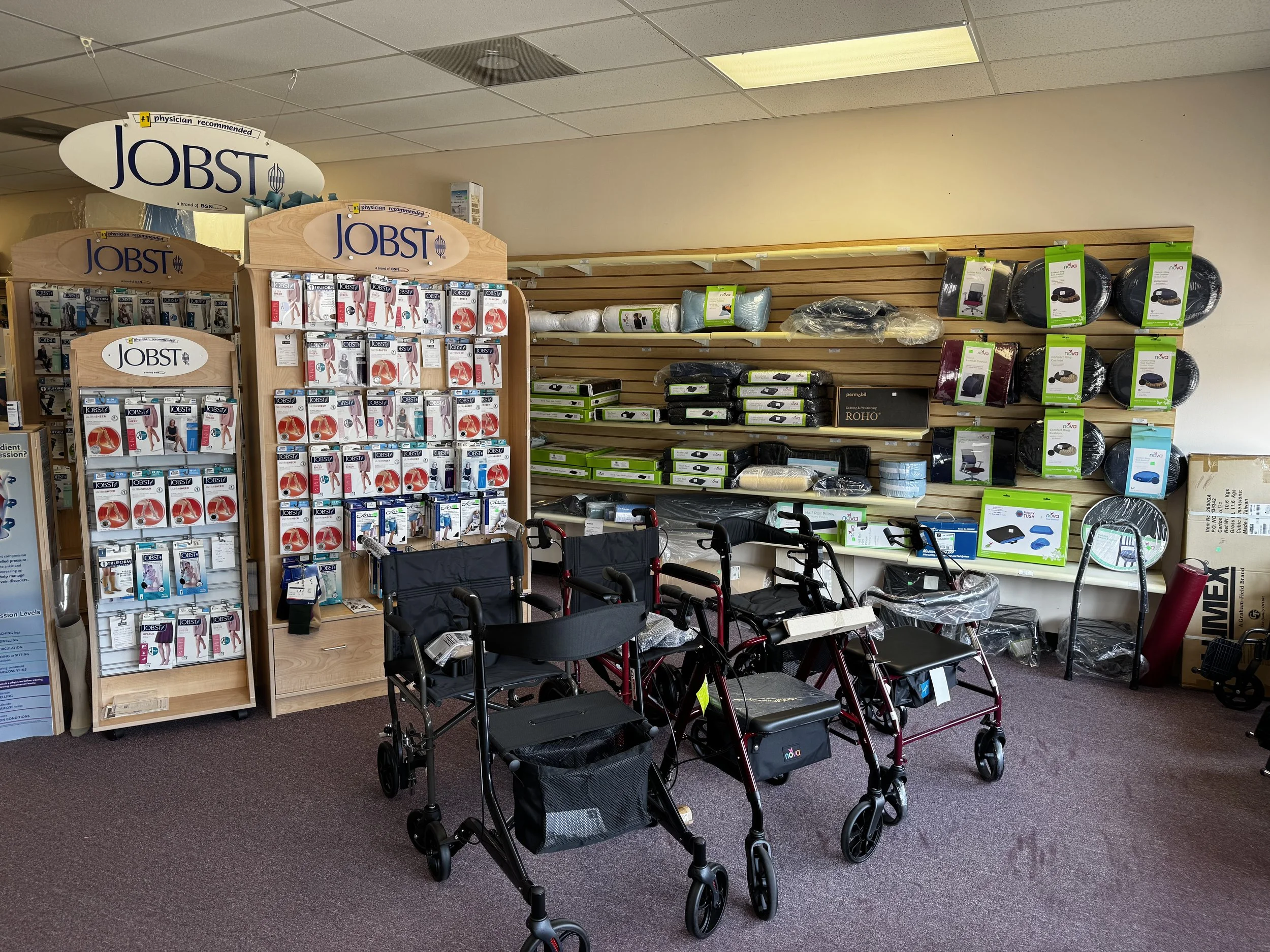 A store aisle with mobility aids and hospital beds on display. There are three rollators in the foreground, and shelves on the wall with cushions, bed frames, and other medical accessories.