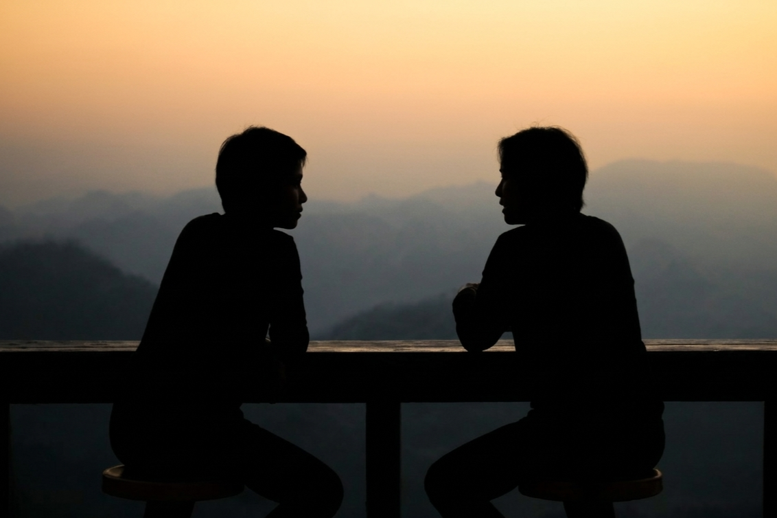 Silhouettes of two people sitting at a table outdoors during sunset with mountains in the background.