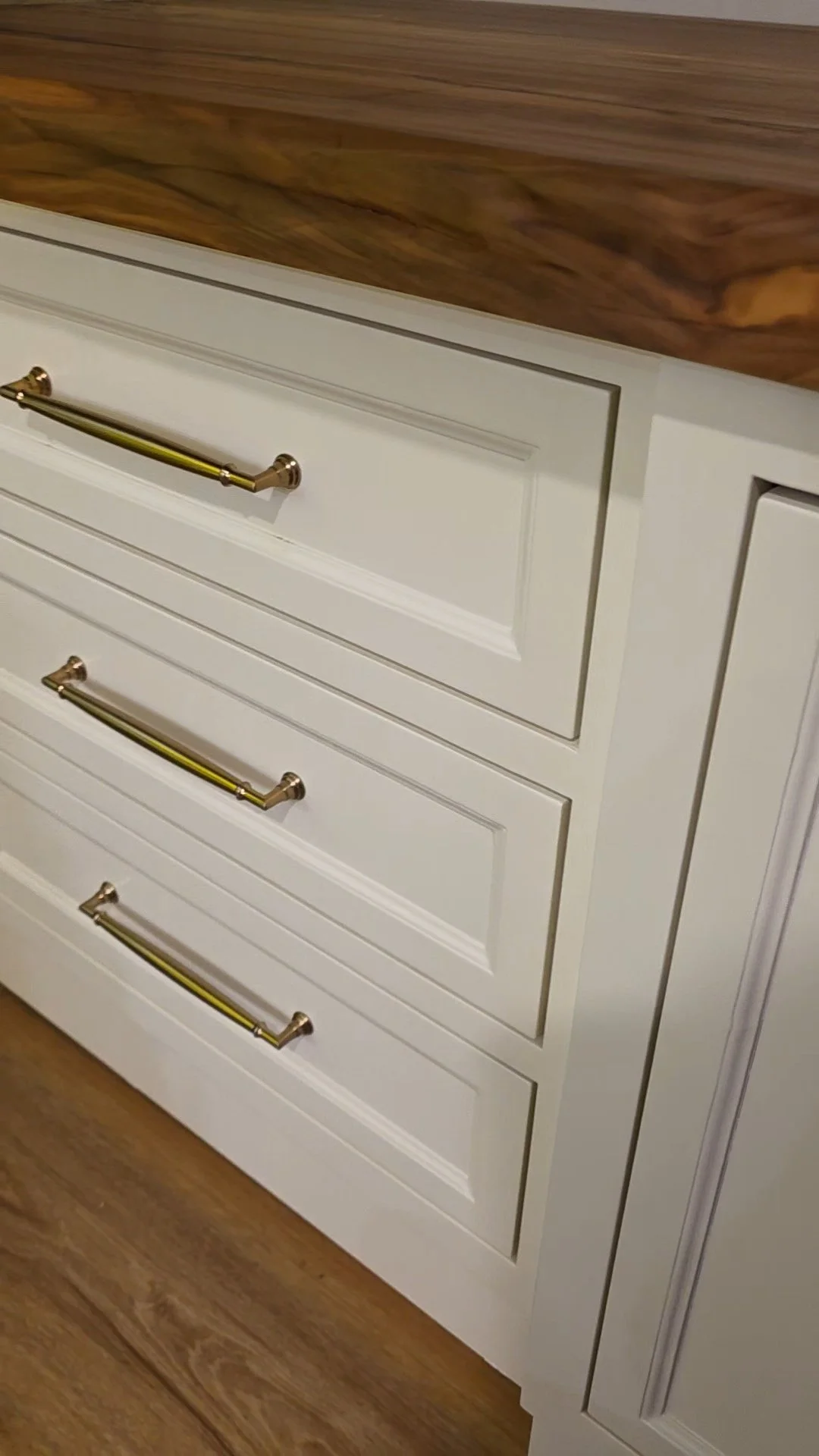 White kitchen cabinet drawers with gold handles, part of a wood countertop, on a wooden floor.