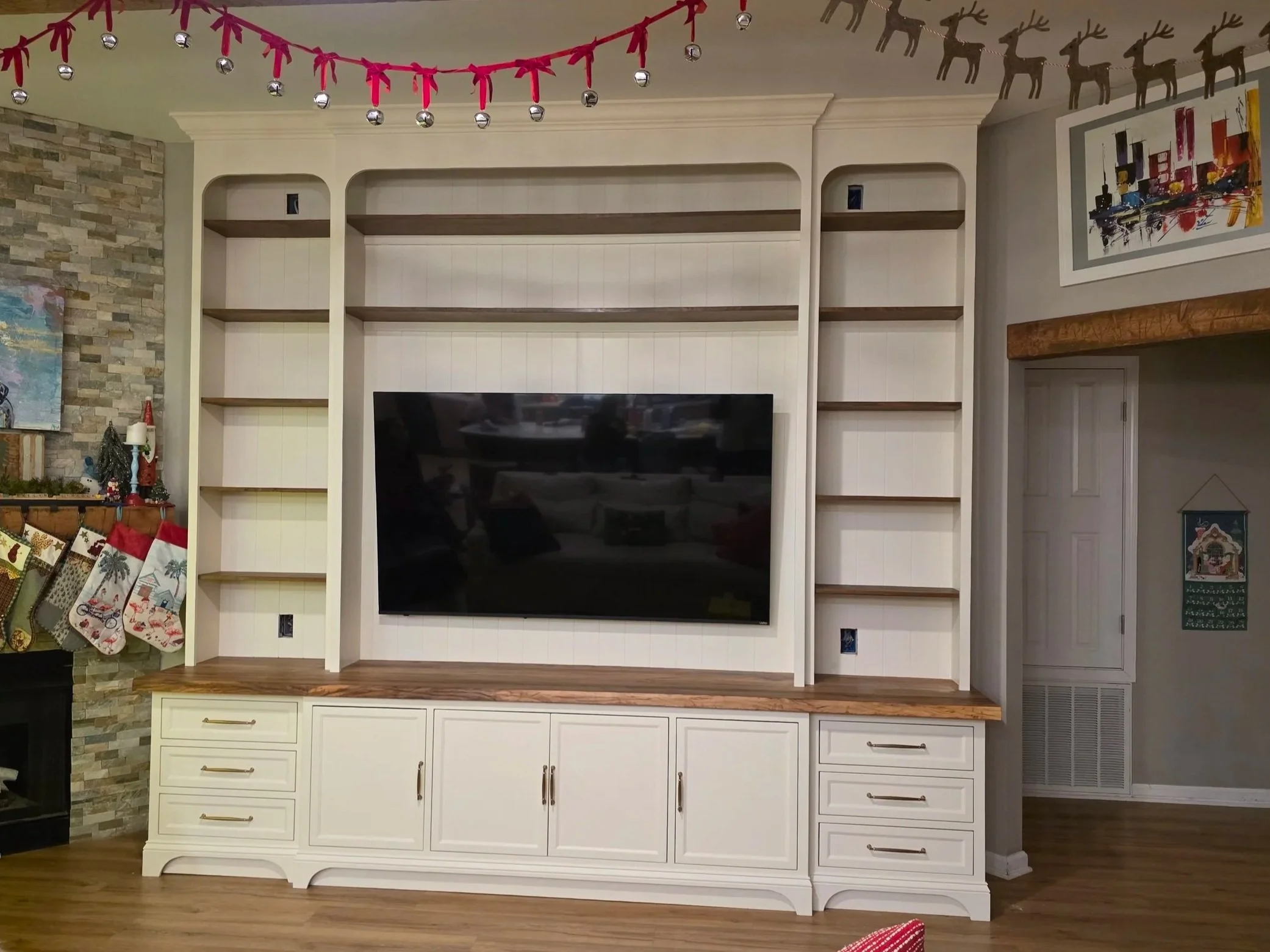 Finished custom built-in entertainment center with shelves and drawers in a living room, decorated for Christmas with stockings hanging on a nearby fireplace and holiday garland across the ceiling.