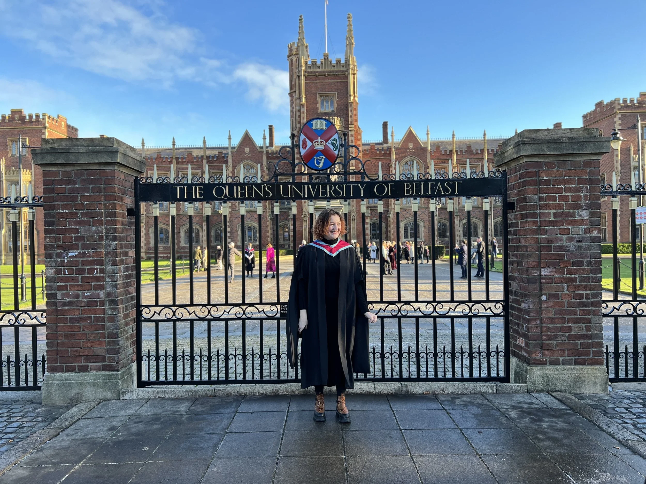 A person with short, curly red hair in a long black robe and red sash stands in front of a gate and seal reading "Queen's University Belfast" with a tall, gothic brick building in the background. They are smiling and looking to the right of the frame