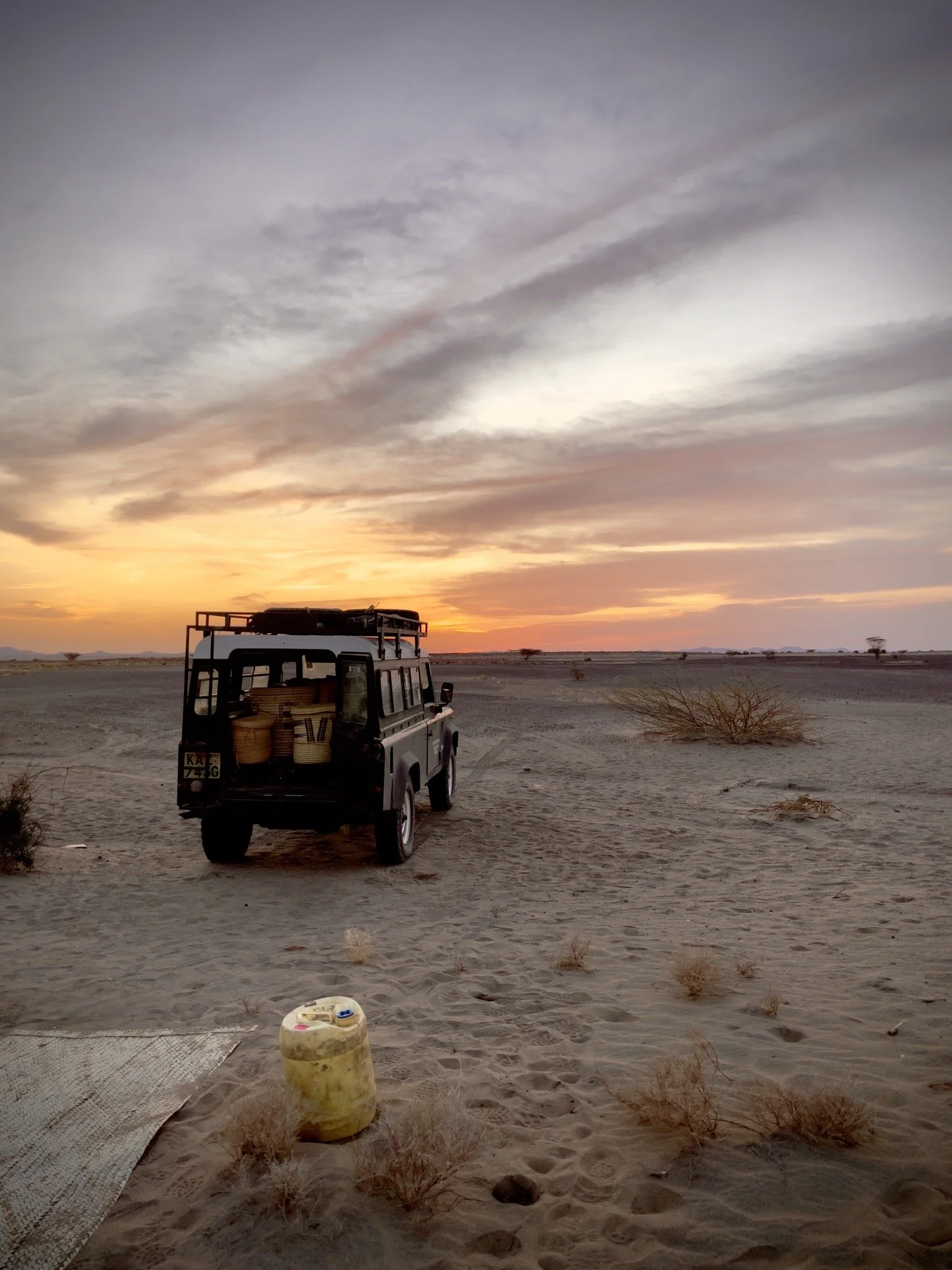 A black off-road vehicle parked in a desert during sunset with a sky filled with clouds, some bushes on the sand, and a yellow container in the foreground.