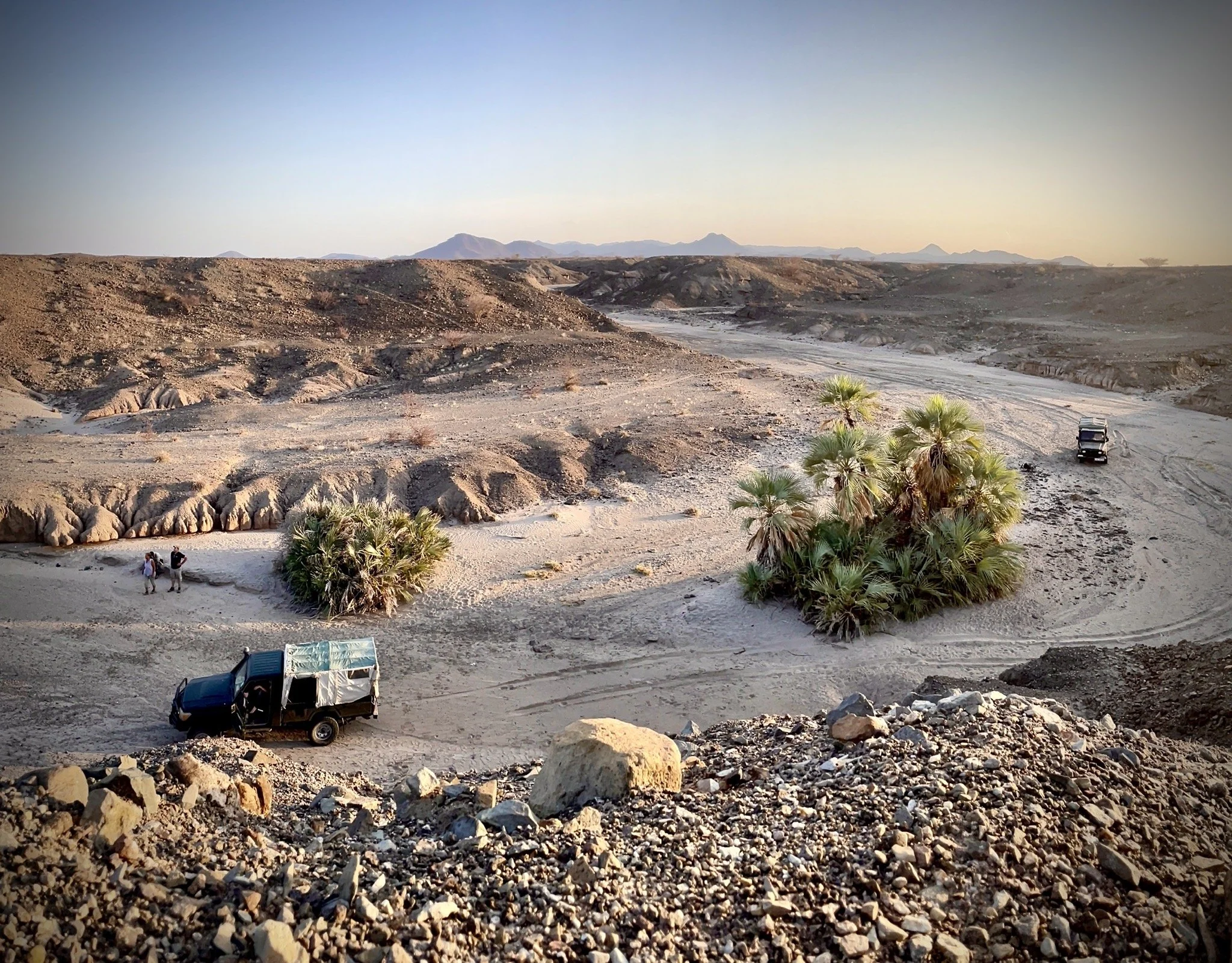 Desert landscape with rocky terrain, sparse vegetation, and palm trees. Two vehicles, one partially visible, parked nearby. Three people standing near the palm trees, mountains in the distant background, and a dirt road or dry riverbed winding throug