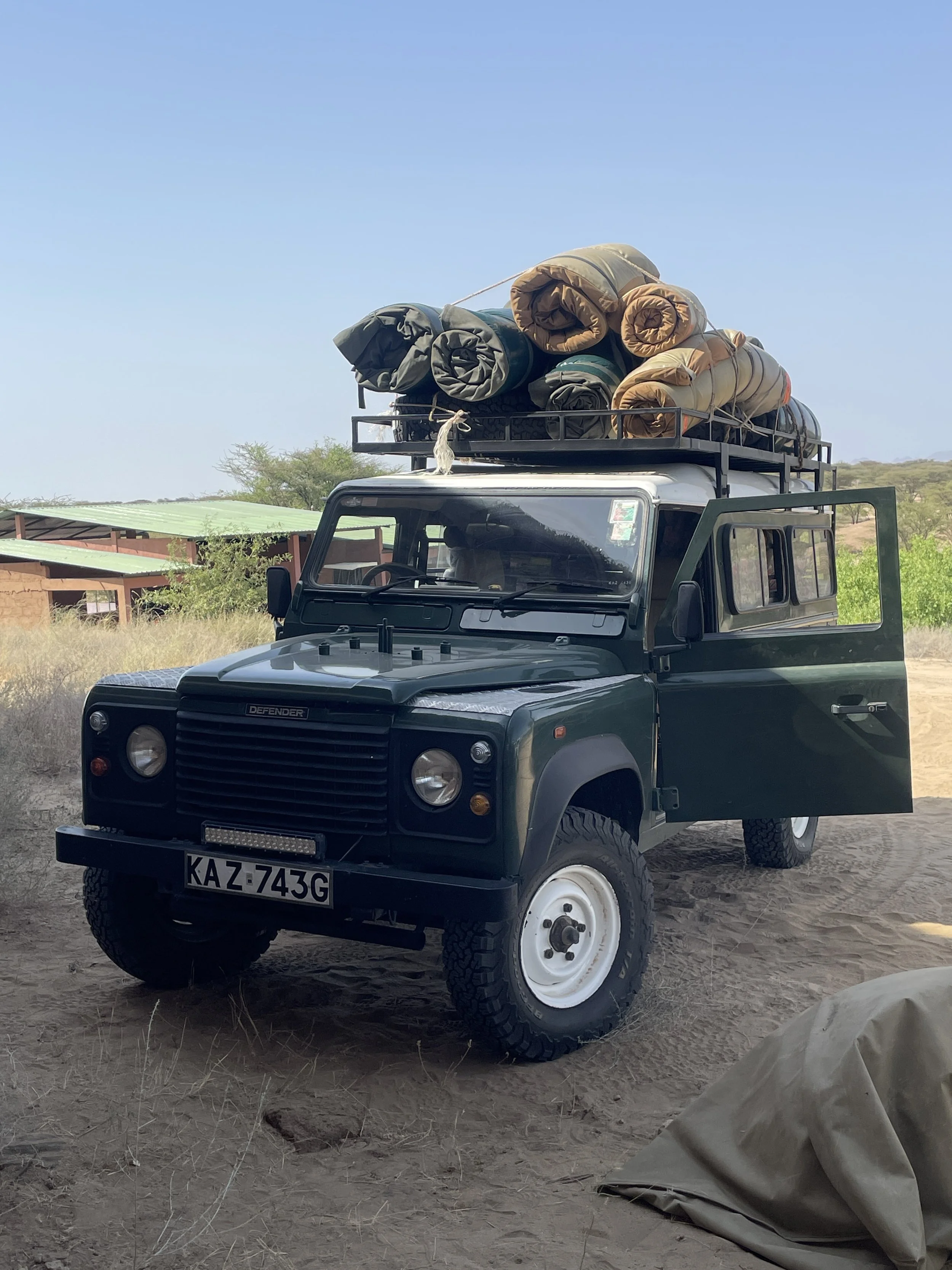 A black Land Rover Defender parked on dirt ground with its front wheel facing camera and the driver's side door open. The roof rack is loaded with rolled camping or sleeping mats. The background shows a building with a green roof and a clear blue sky