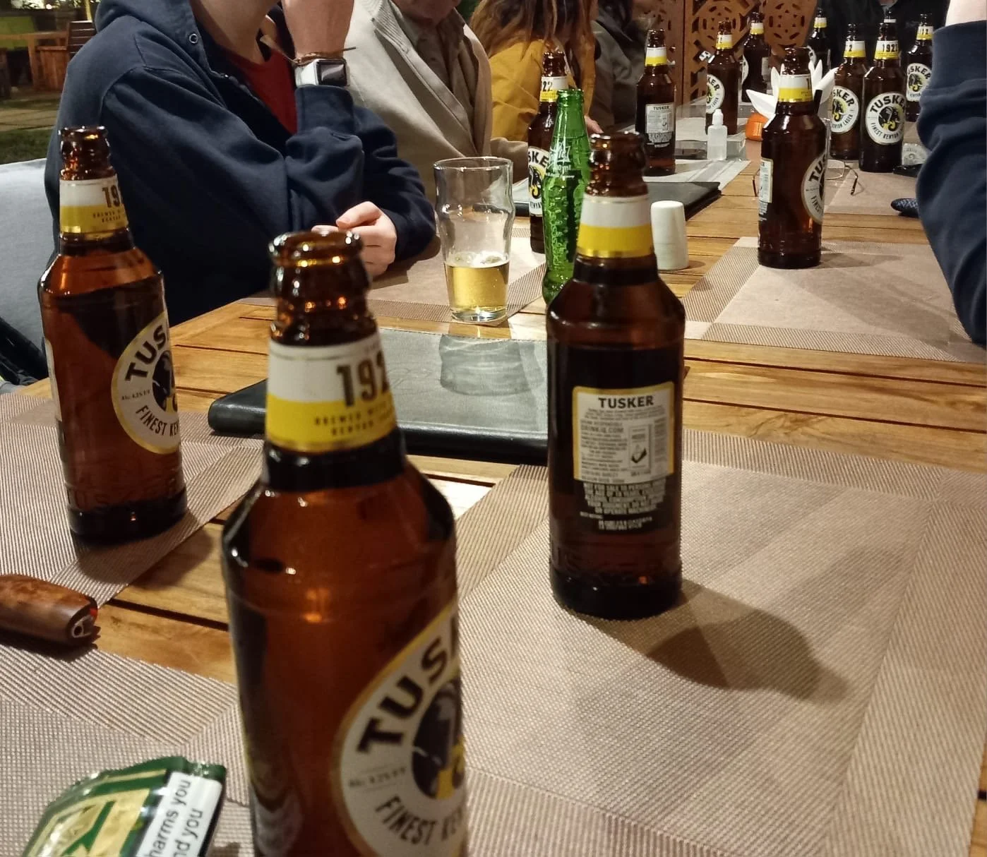 A group of people sitting around a wooden table outdoors, with beer bottles, a glass of beer, and some condiments visible on the table.