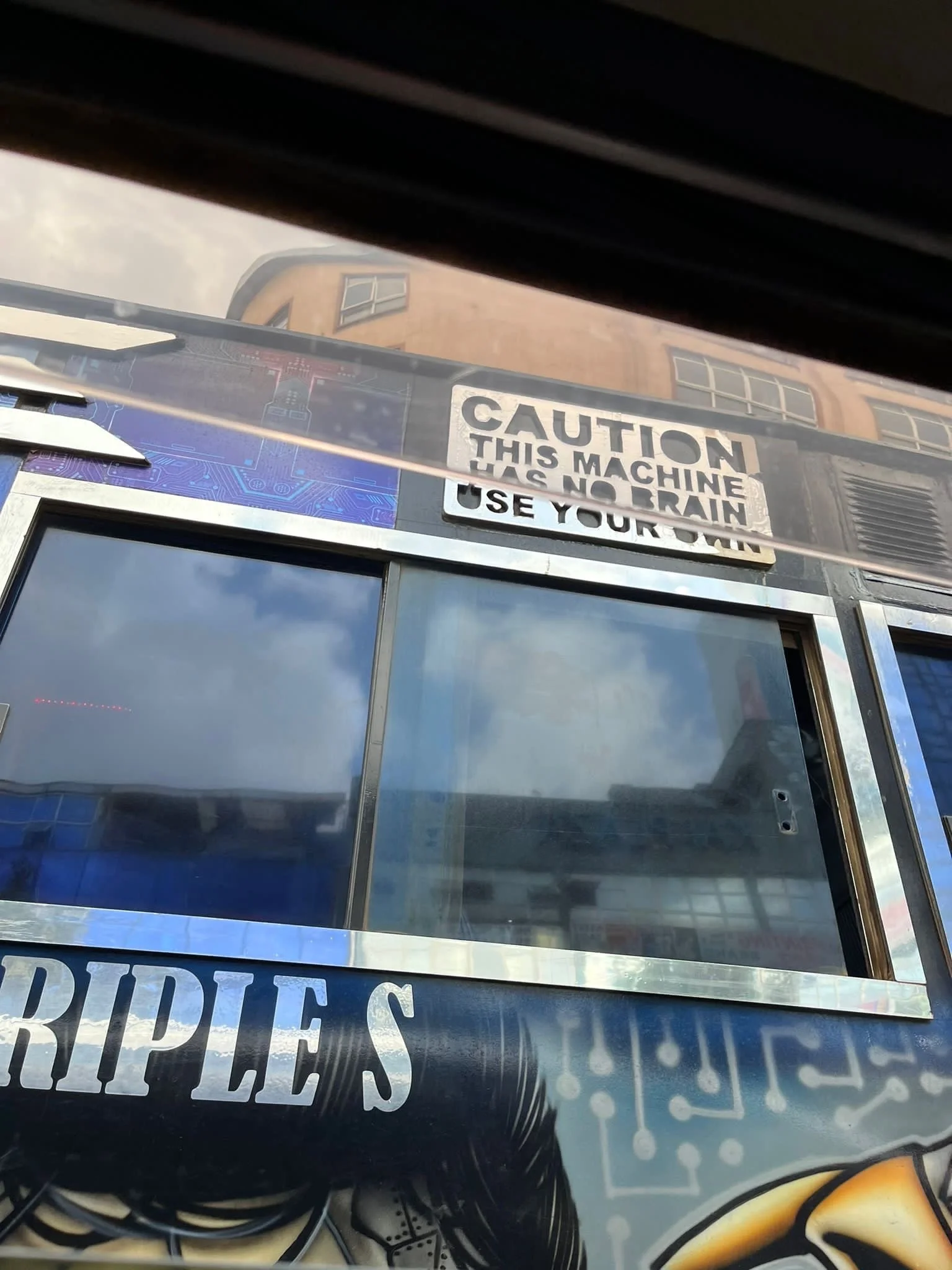 Close-up of a vending machine with a caution sign that reads "Caution, this machine has no brain. Use your head." The machine has reflective glass and some wires visible at the bottom, with a building reflected in the glass.
