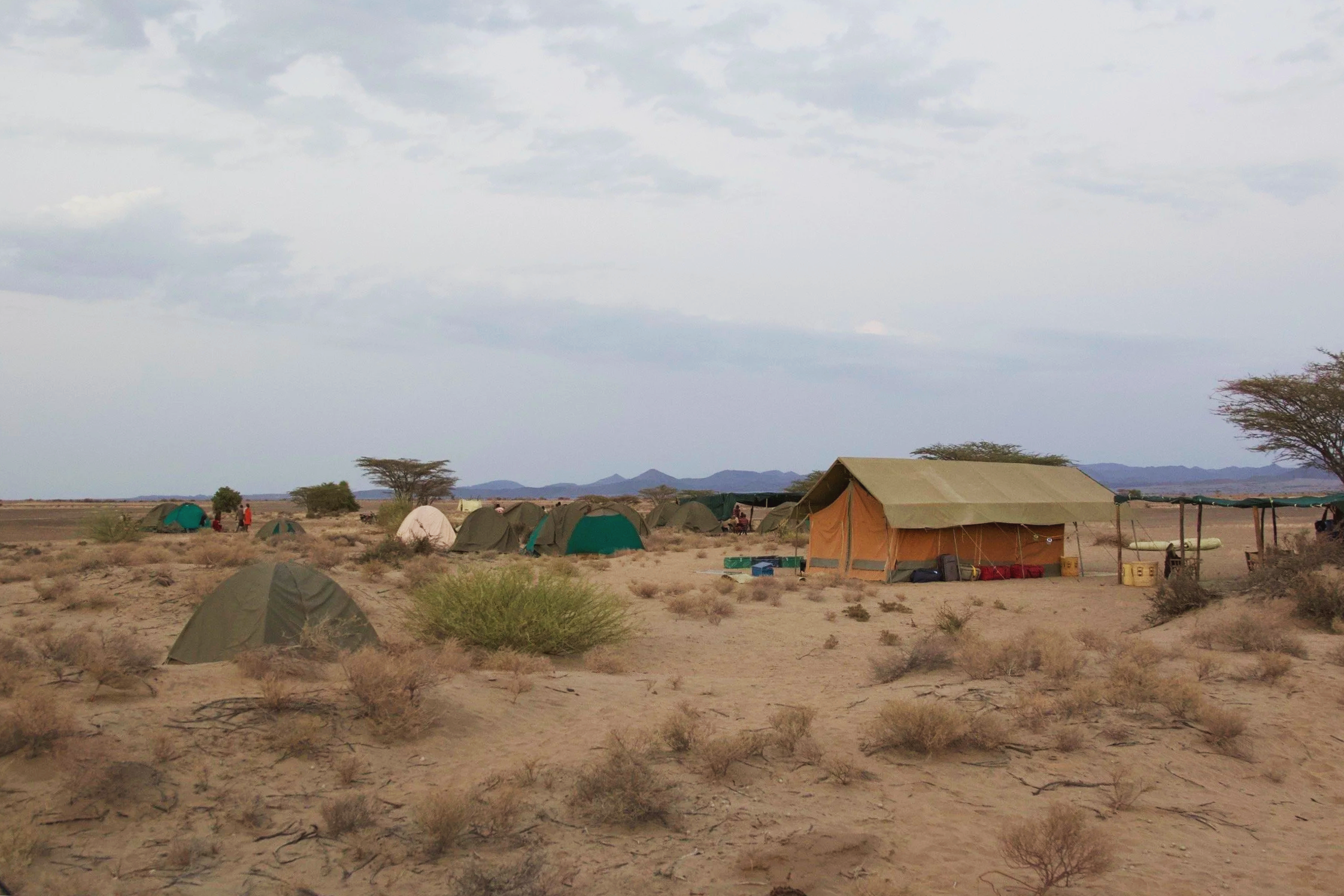 A desert campsite with multiple tents and a large shelter, surrounded by sparse vegetation, under a cloudy sky.