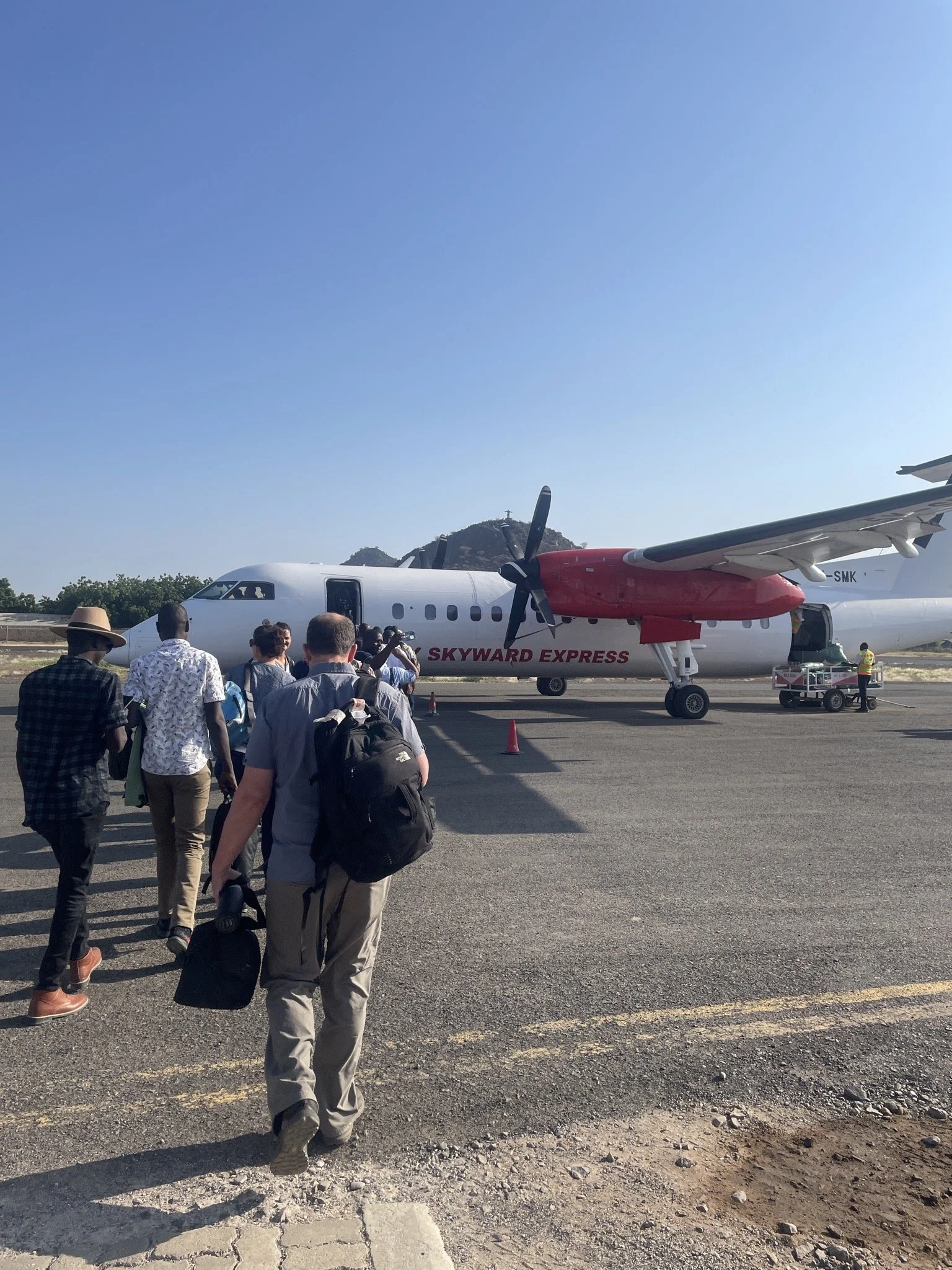 Group of travelers walking towards a small commercial airplane on the tarmac, with a clear blue sky and a mountain in the background.
