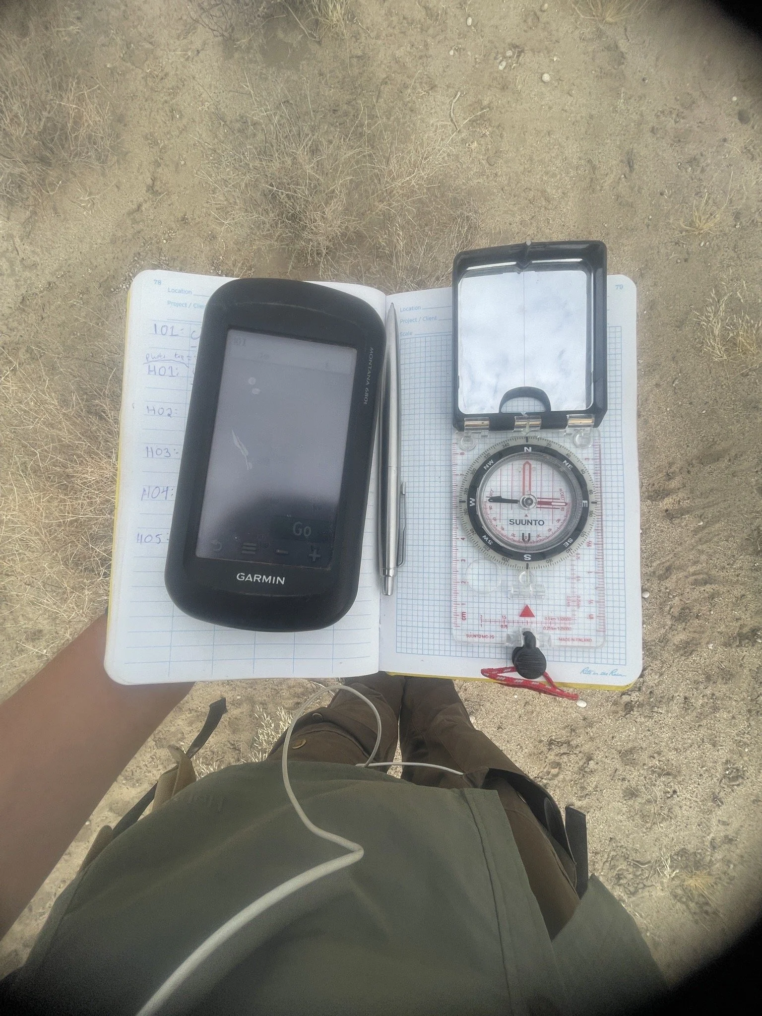 A handheld GPS device, a compass with a mirror, a pen, and a notebook with handwritten notes are placed on a grid paper in a desert-like environment with dry grass and dirt.