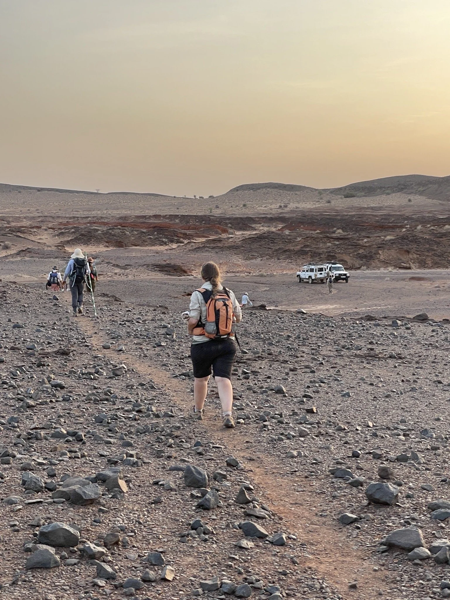 Group of people hiking in a desert landscape during sunset, with two white off-road vehicles parked in the distance.